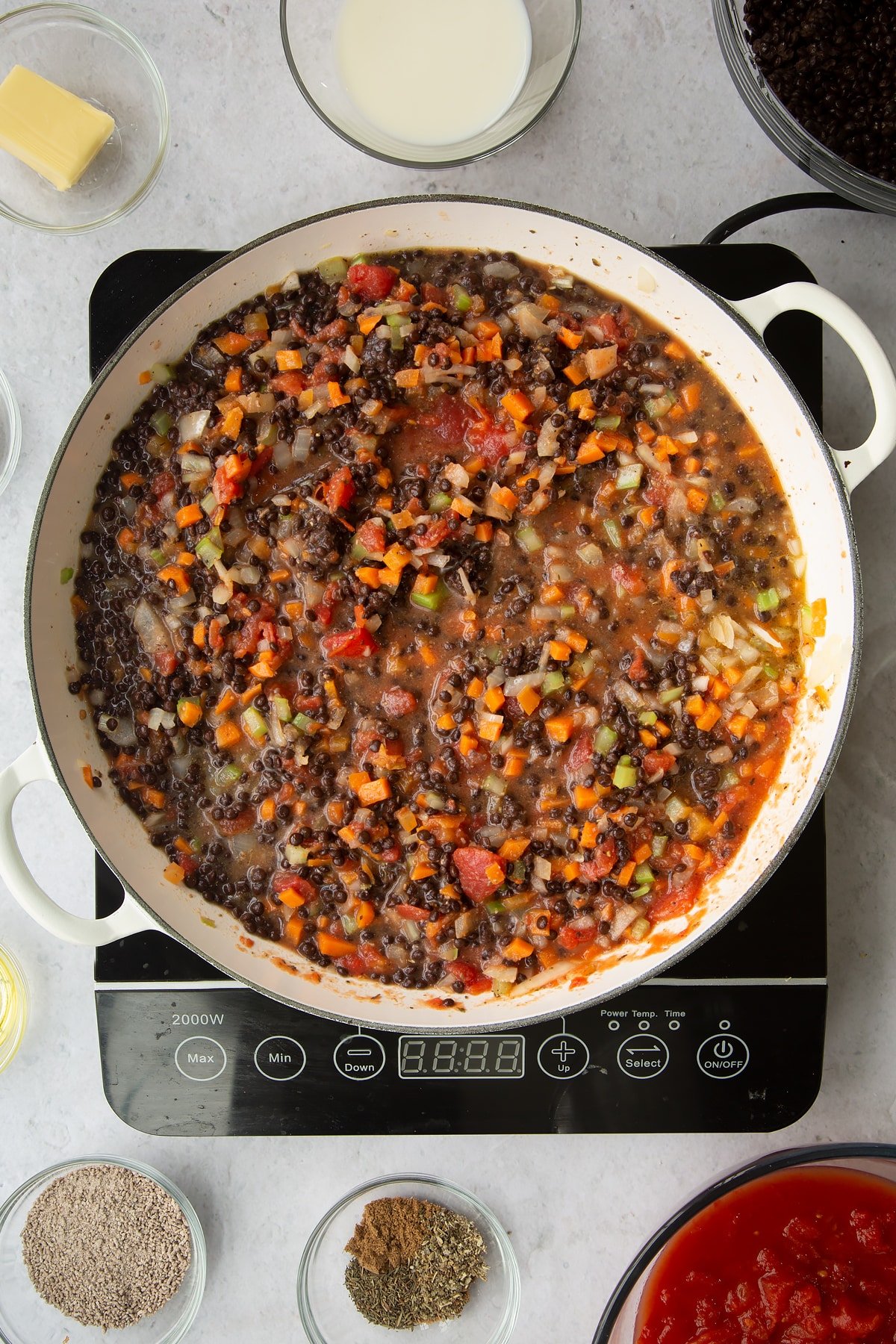 a large frying pan on an induction hob with mixed vegetables, brown lentils and tinned tomatos mixed together.