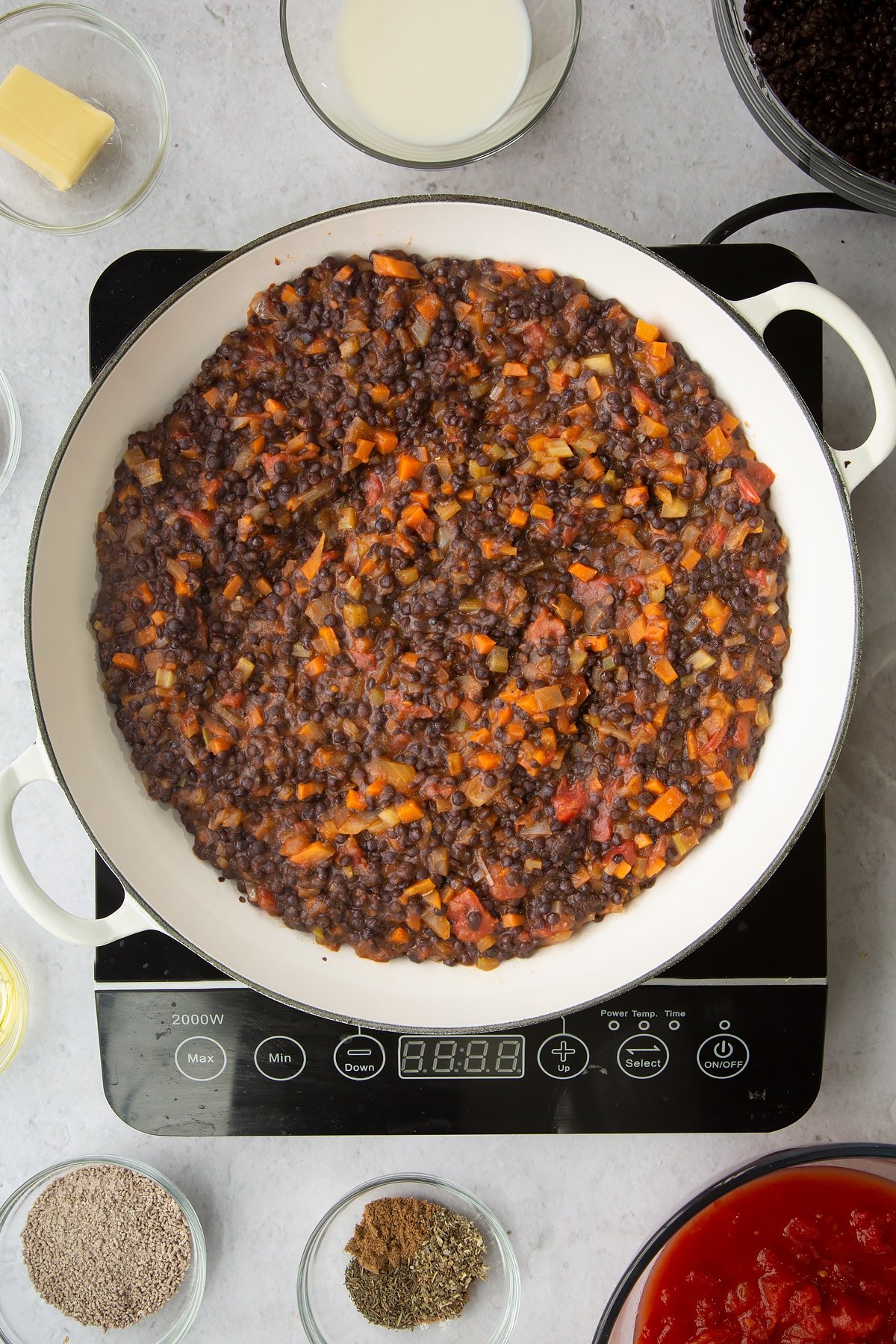 a large frying pan on an induction hob with mixed vegetables, brown lentils and tinned tomatos mixed together.