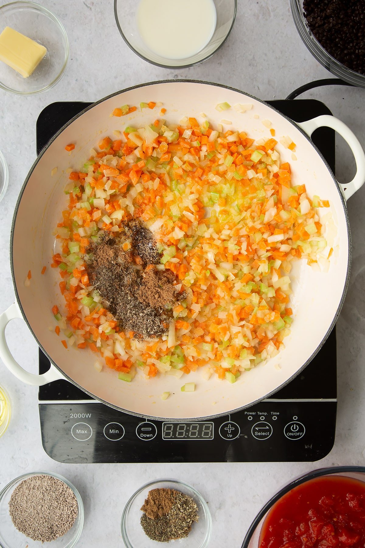 a large frying pan on an induction hob with chopped onion, celery, carrot, garlic with added herbs.