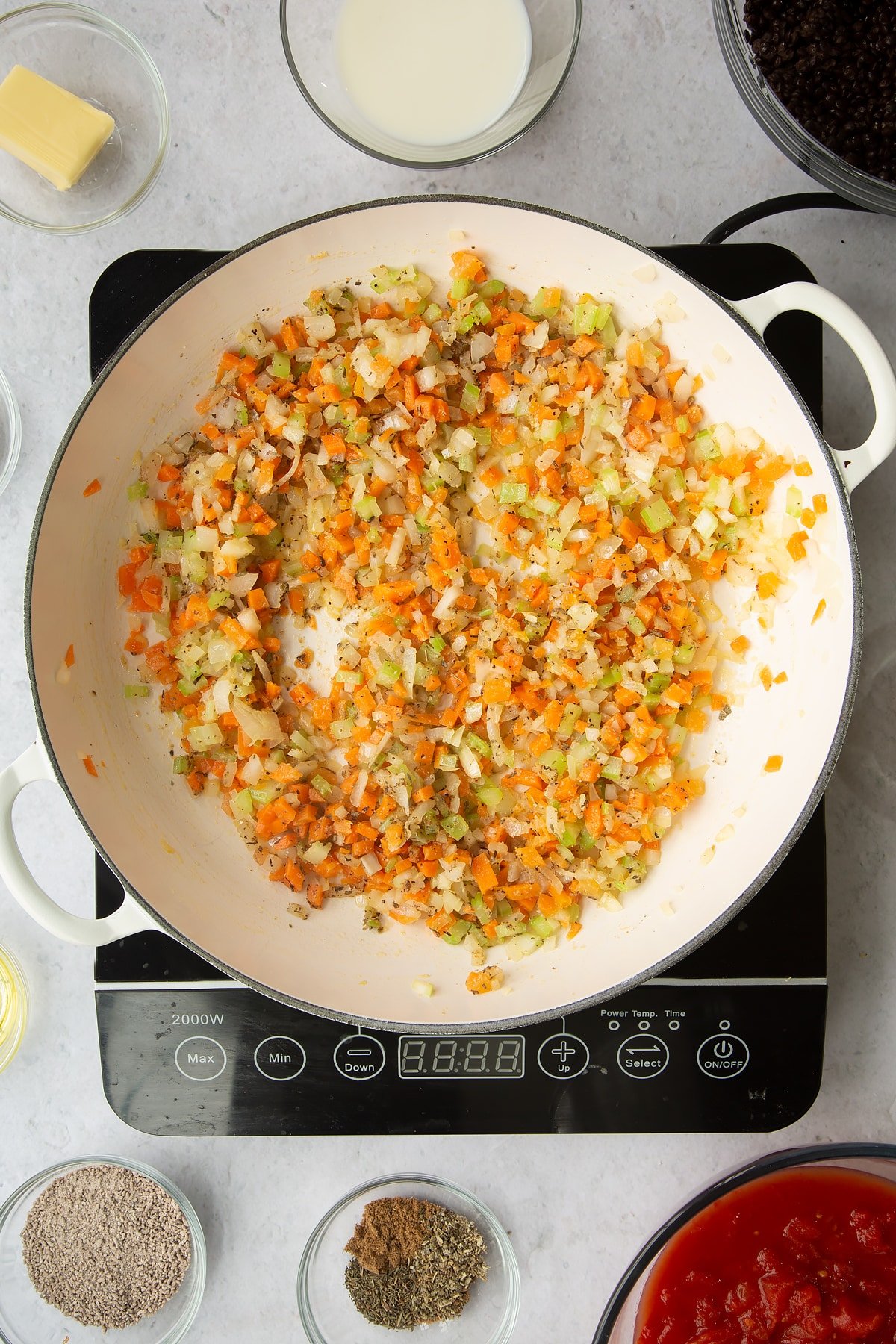 a large frying pan on an induction hob with chopped onion, celery, carrot, garlic with added herbs.