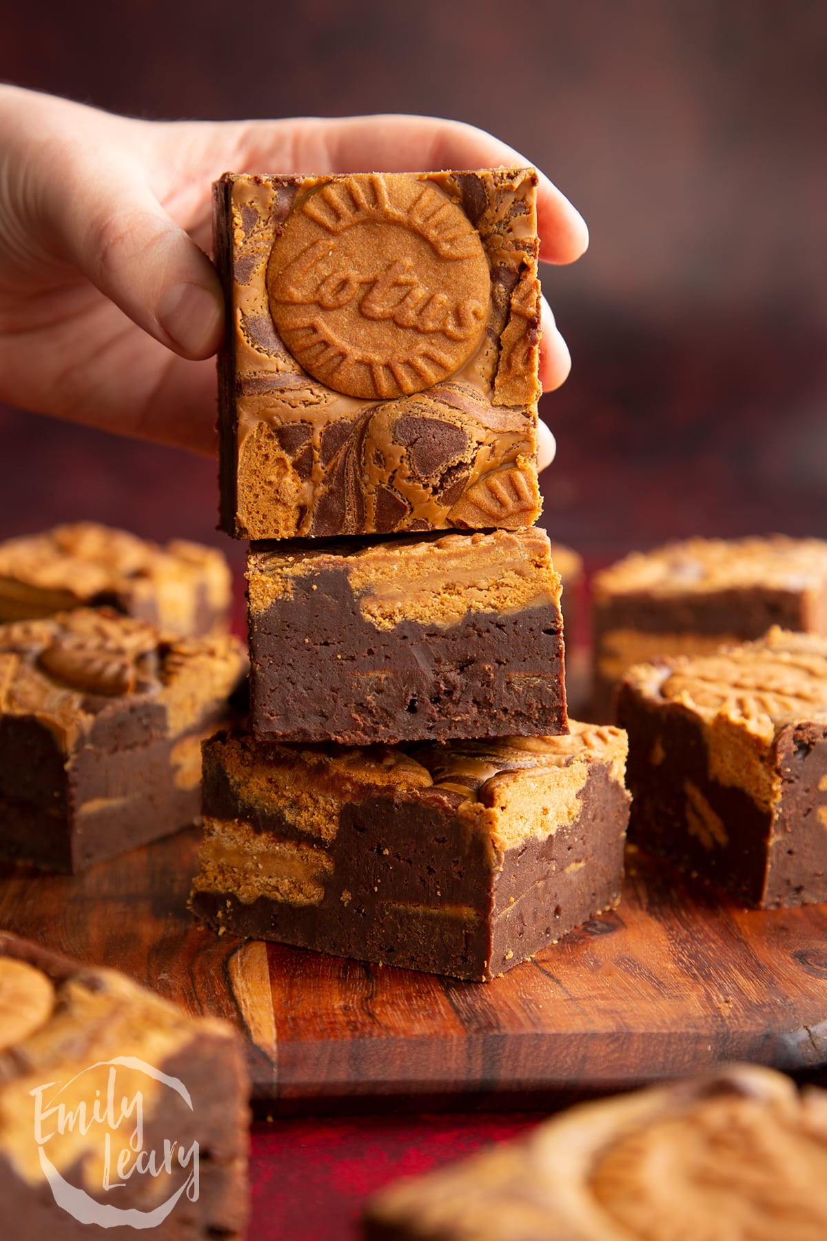 a hand holding a Biscoff brownie on a stack of more Biscoff brownies on a wooden chopping board.
