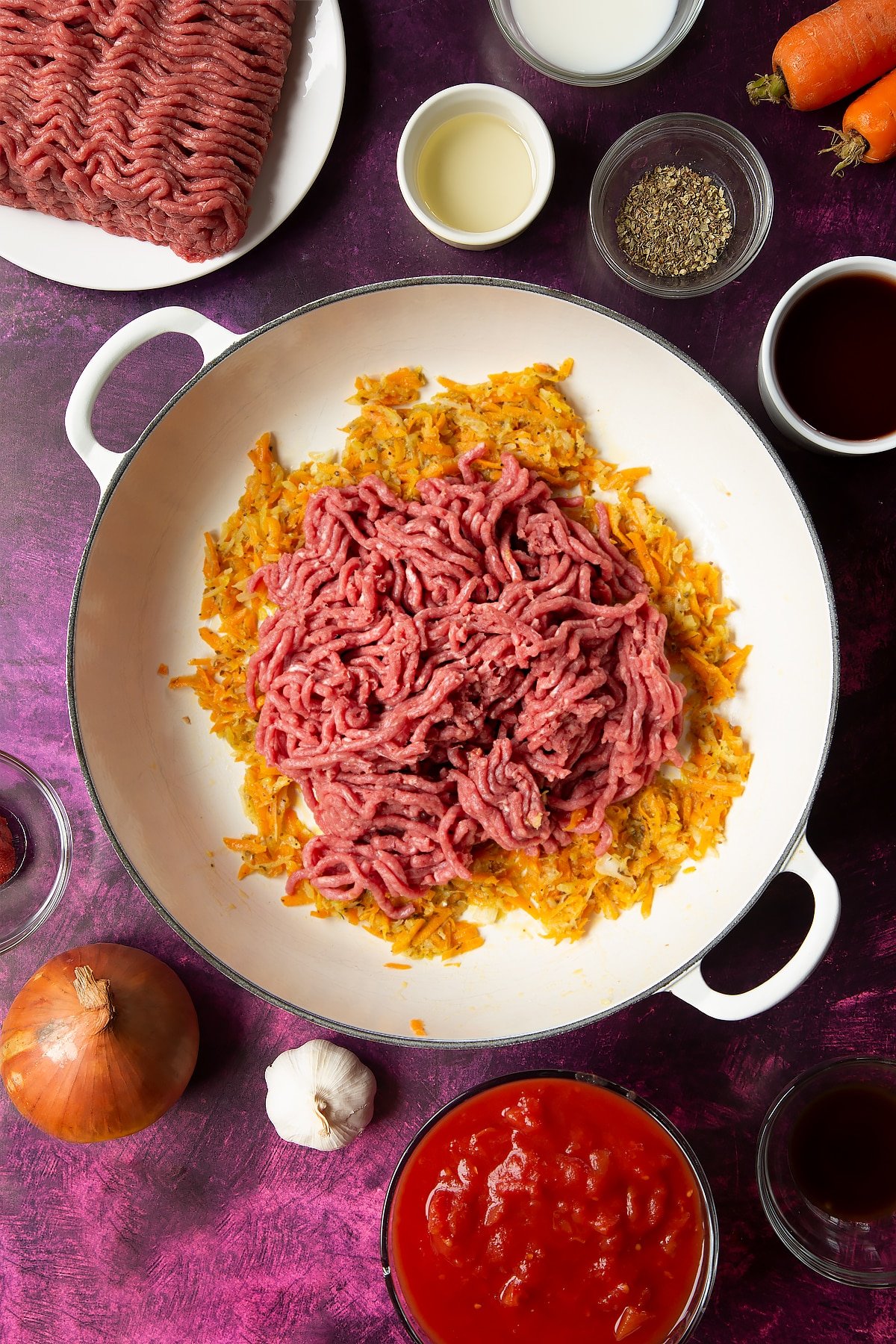 cooked carrot and onion mix in a large white frying pan topped with raw beef mince.
