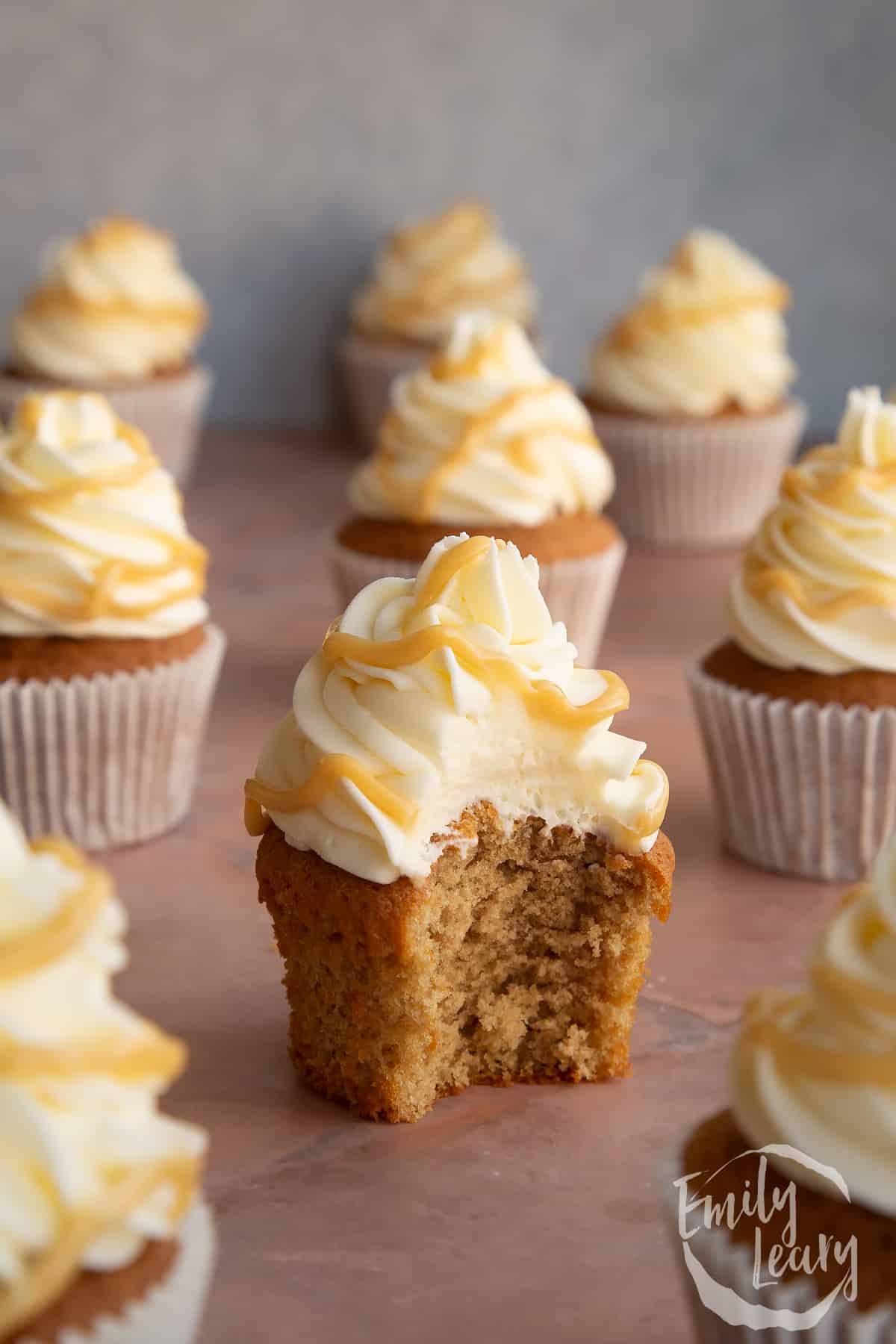 a Caramel latte cupcake with a bite missing from the side surrounded by more cupcakes.
