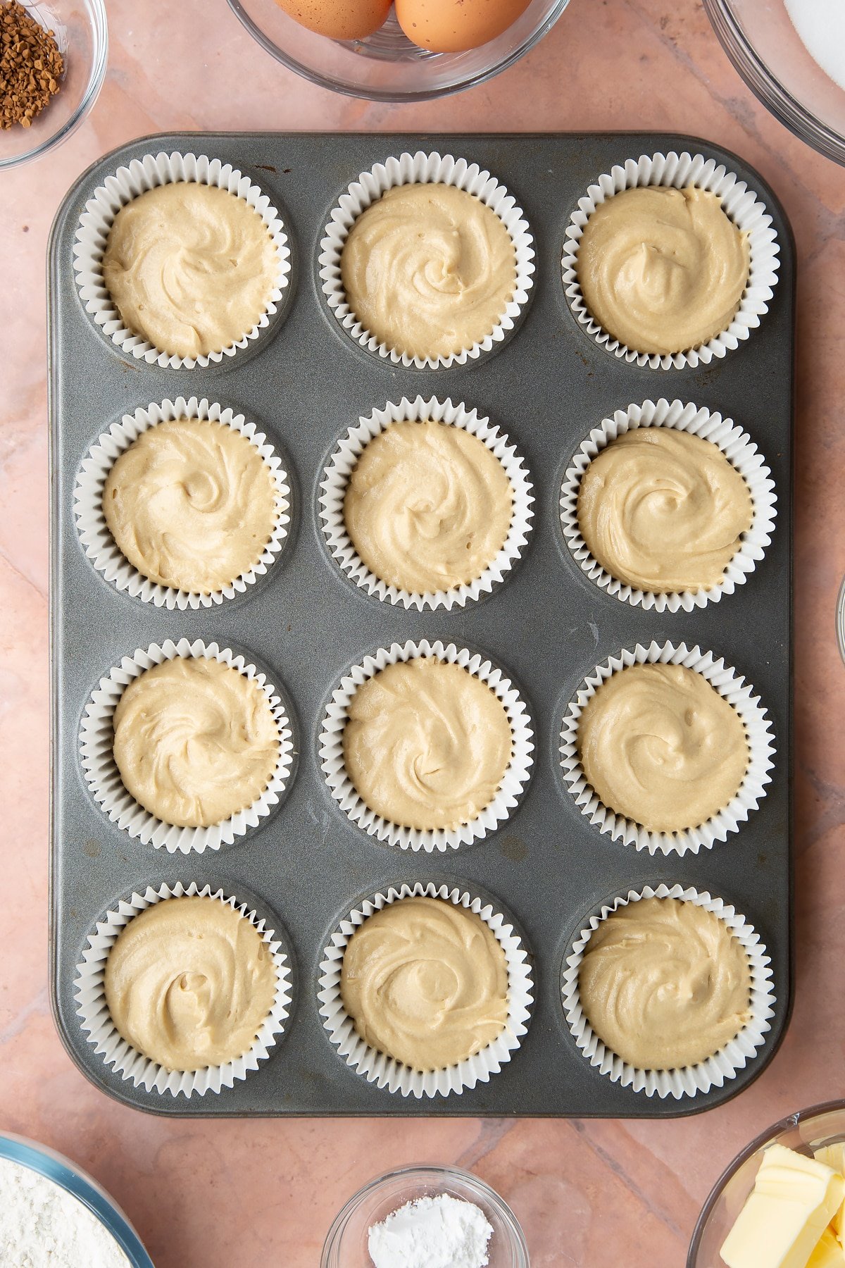 coffee cake batter in white bun cases in a bun tin on a pink background.