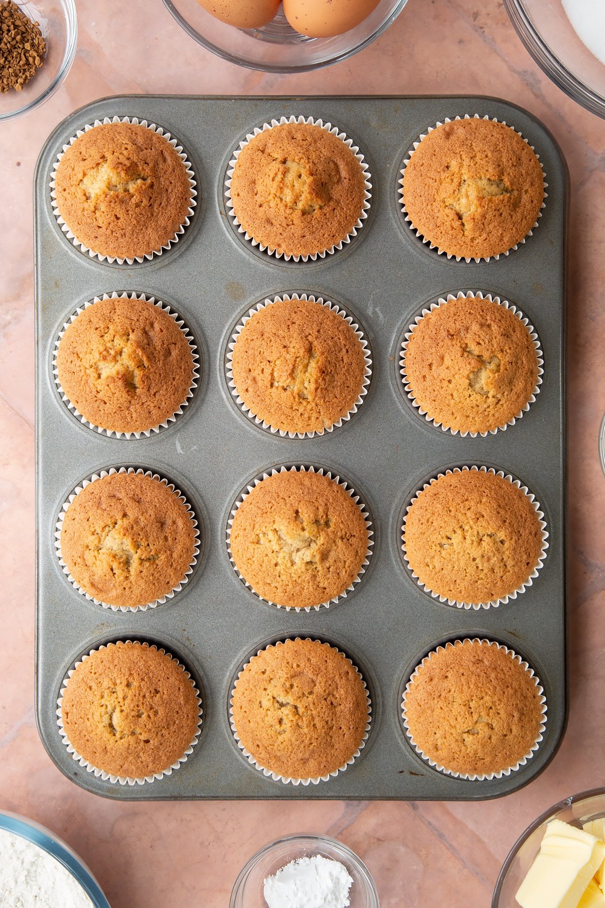 baked coffee cake cupcakes in white bun cases in a bun tin on a pink background.