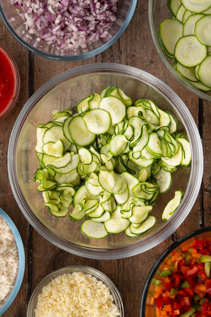thinly sliced courgette drizzled in oil in a clear bowl with ingredients around the edges.