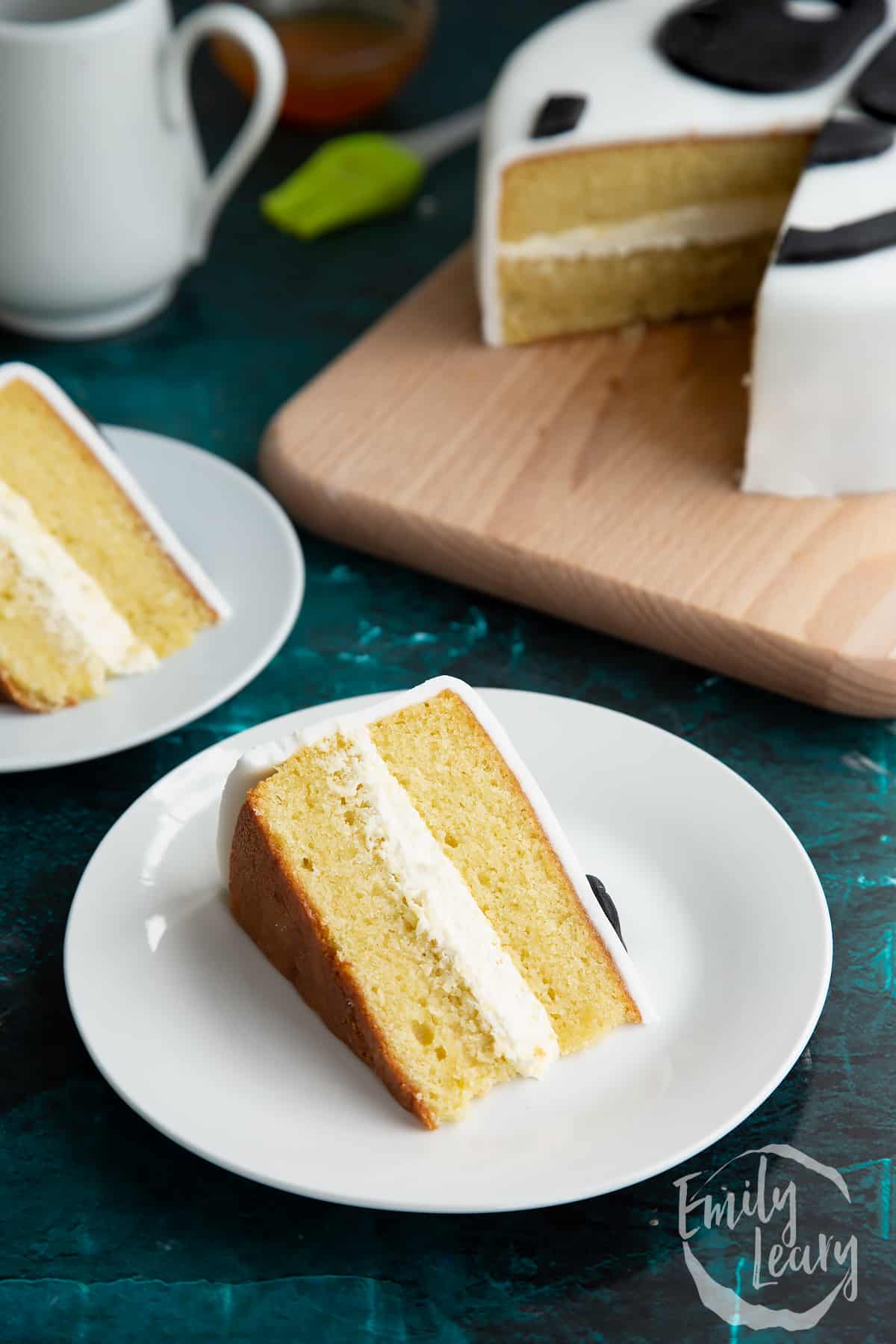 a slice of vanilla cake with buttercream icing on a white plate with the full cake in the background.