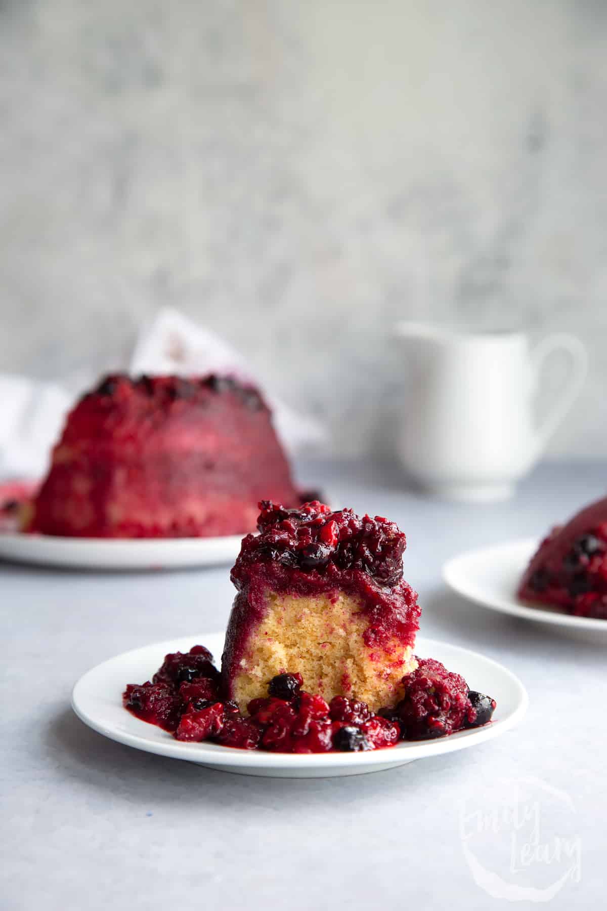 a slice of summer fruit pudding on a white plate with the full pudding in the background.