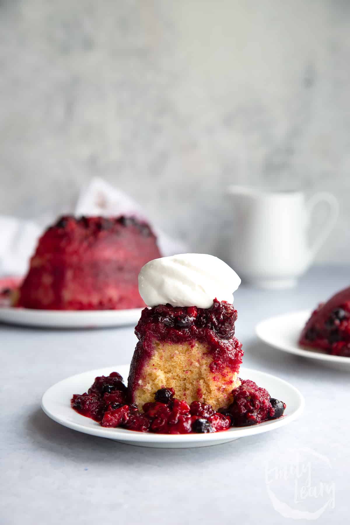 a slice of summer fruit pudding on a white plate topped with whipped cream with the full pudding in the background.