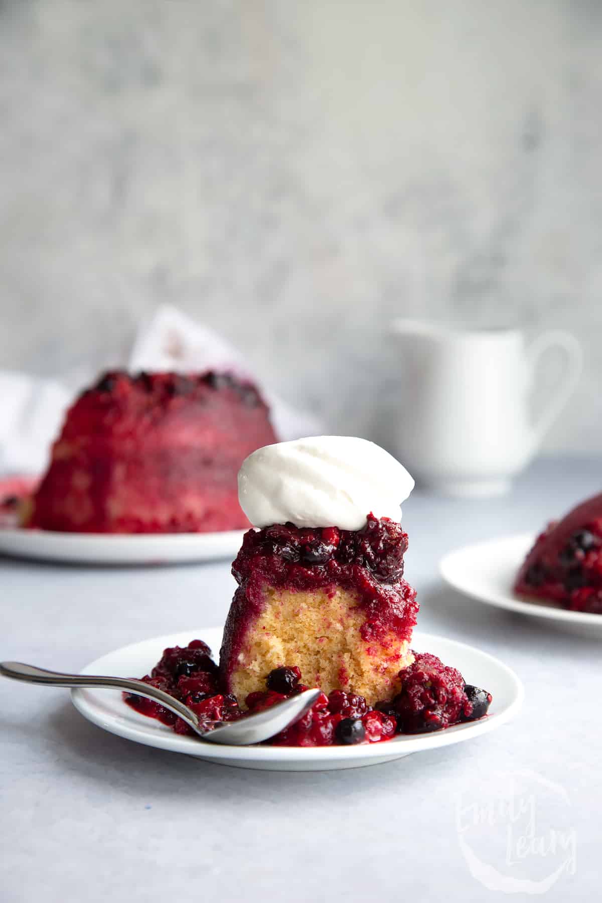 a slice of Summer fruit sponge pudding on a white plate with a spoon on the side topped with whipped cream.