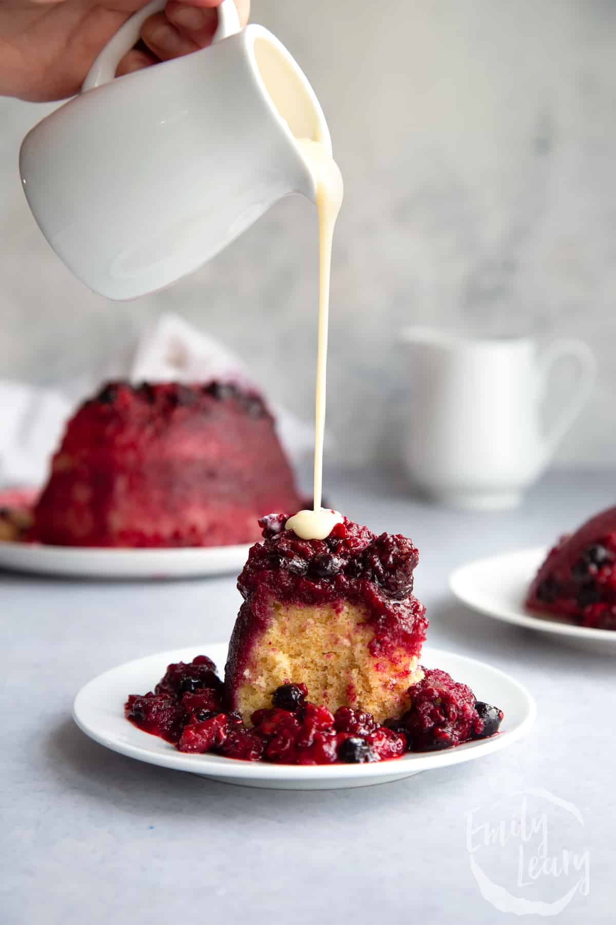 a slice of summer fruit pudding on a white plate with a hand pouring over hot custard.