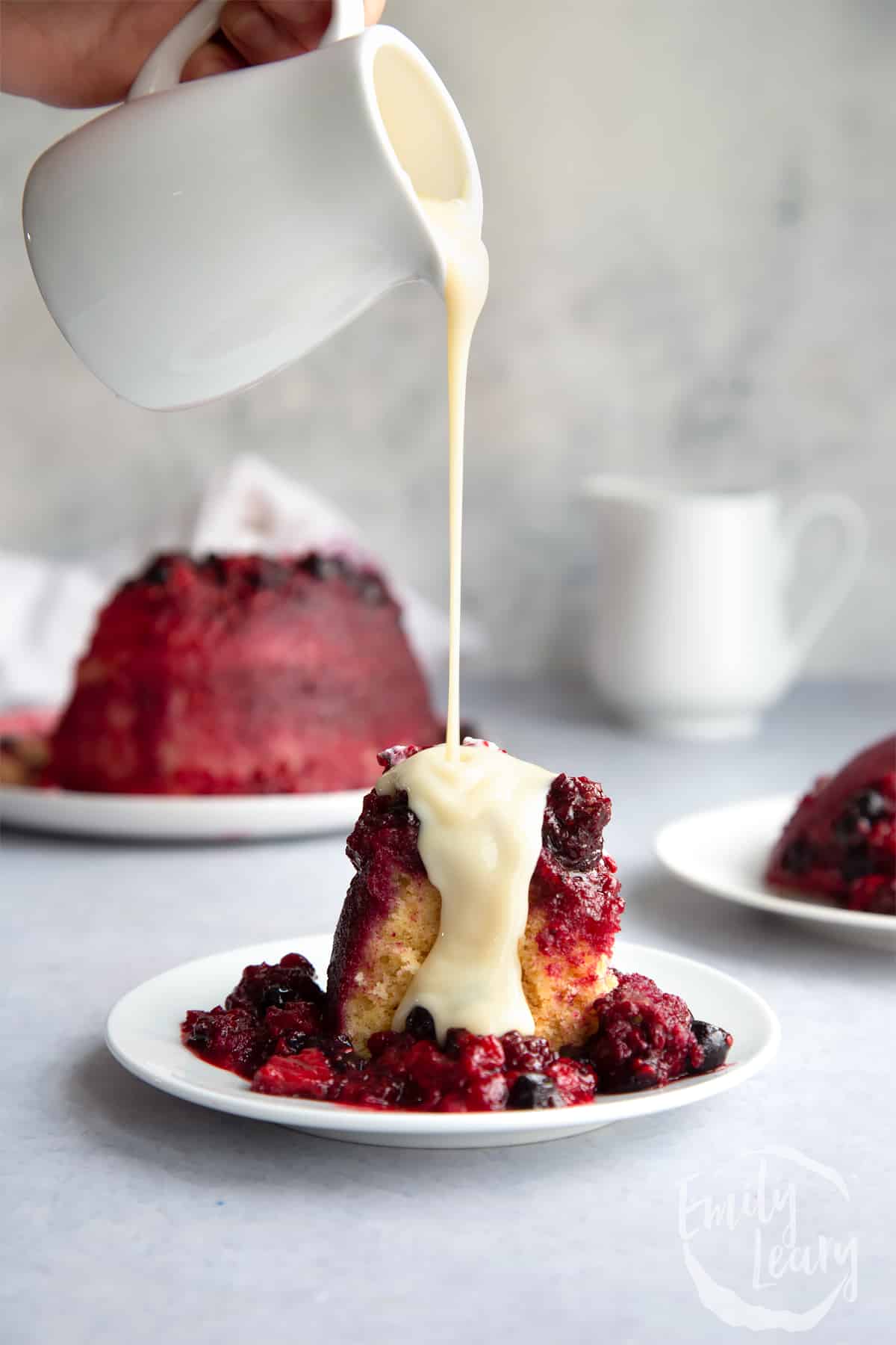 a slice of Summer fruit sponge pudding on a white plate with a spoon on the side with a hand pouring over custard.