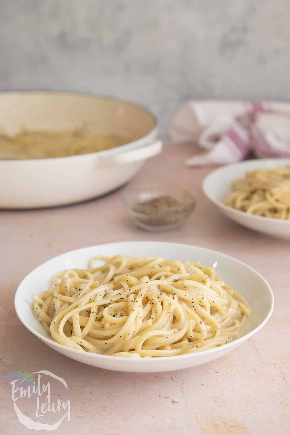 a white bowl of Linguine cacio e pepe on a light surface with a white skillet in the background.