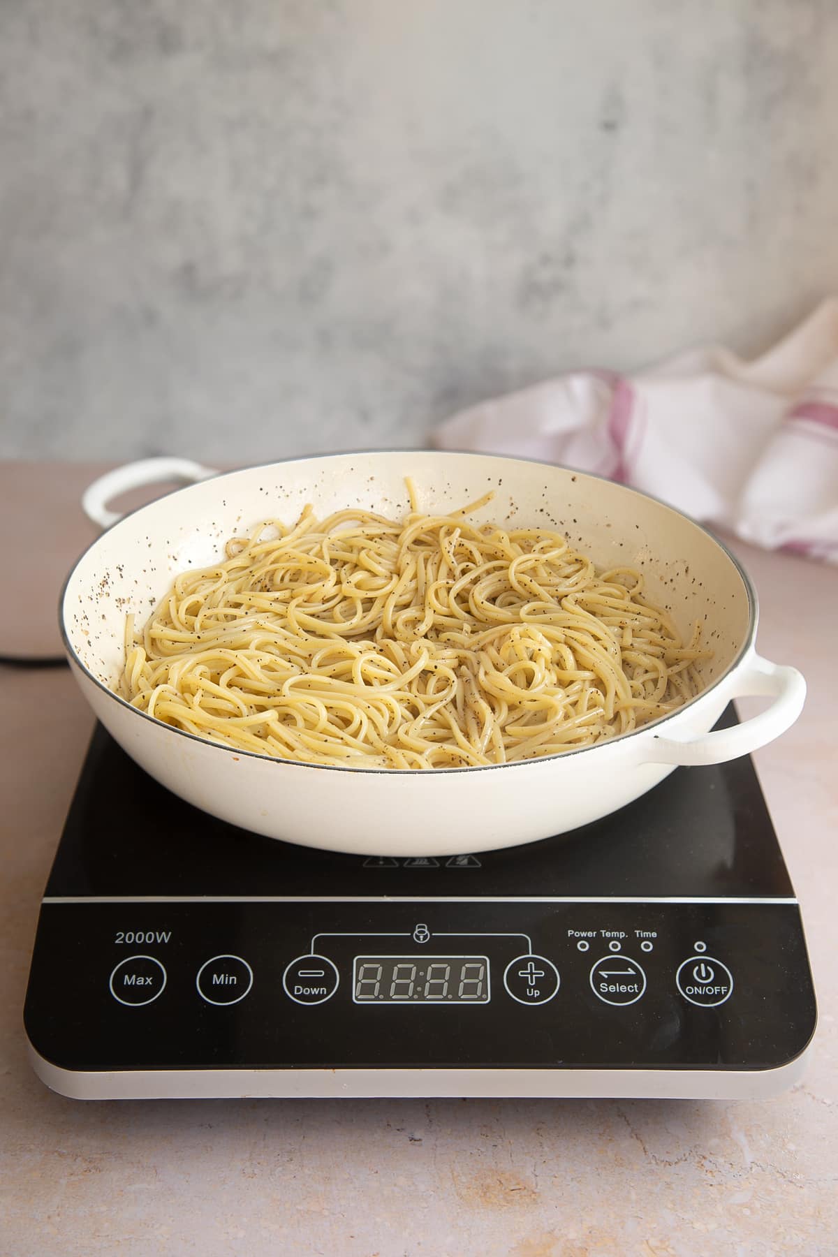 a white skillet pan with oil, pepper and cooked linguine mixed together in a on a induction hob.