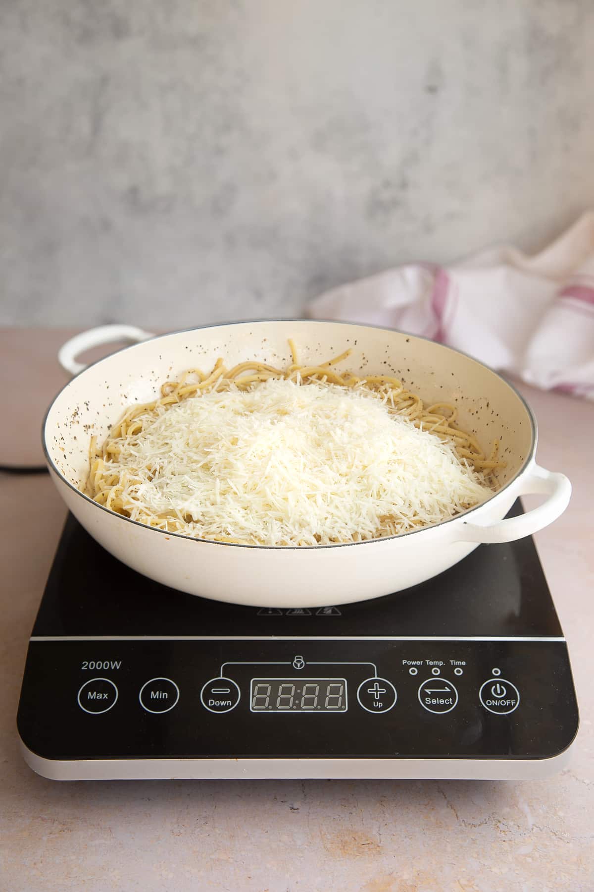 a white skillet pan with oil, pepper and cooked linguine mixed together topped with grated cheese on a induction hob.