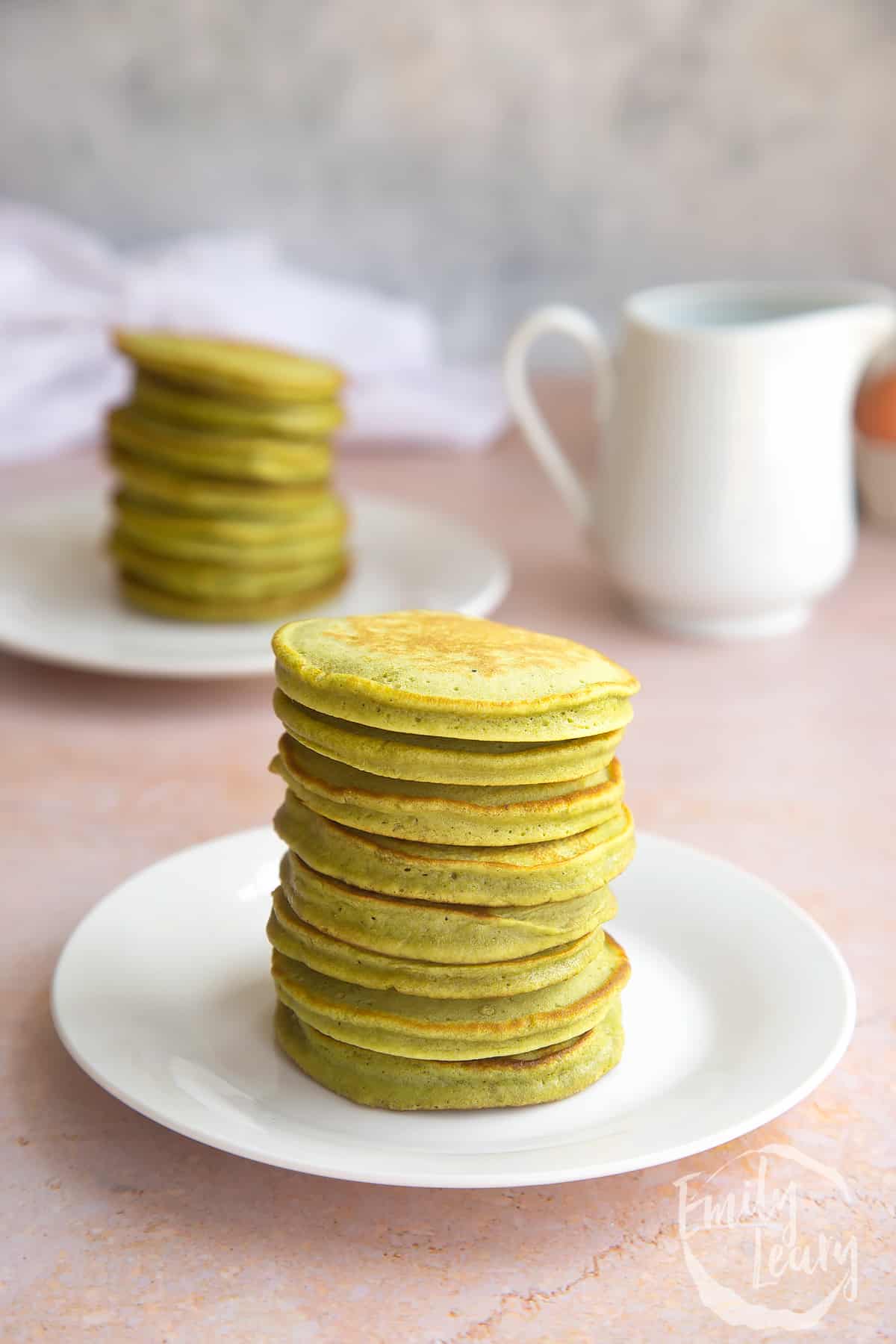 a stack of matcha pancakes on a white plate with more matcha pancakes in the background.