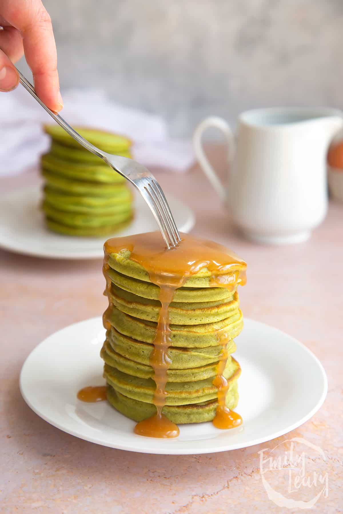a stack of matcha pancakes on a white plate covered in caramel sauce with a fork pointing to the top pancake.