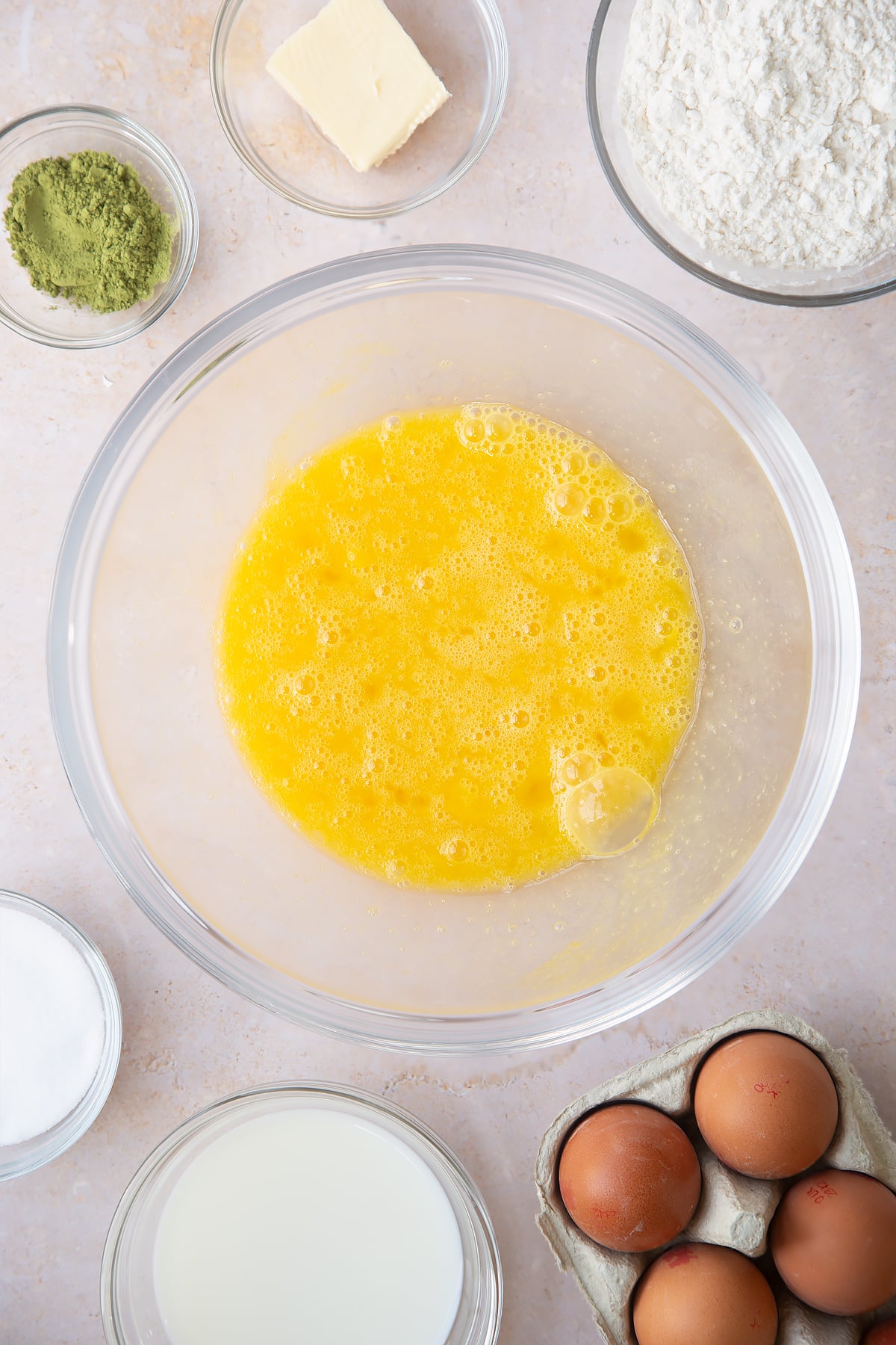 whisked eggs and sugar in a large clear bowl surrounded by ingredients.