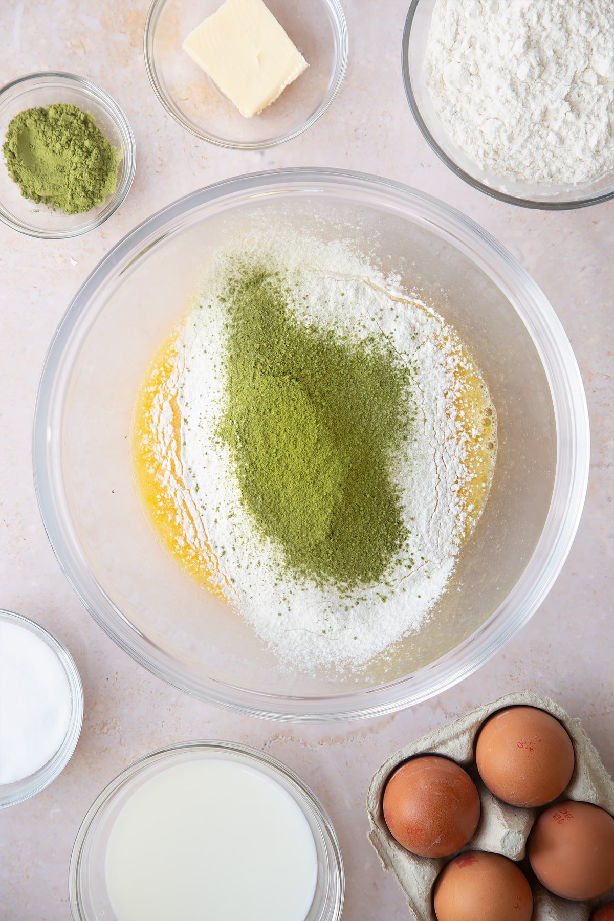 whisked eggs and sugar in a large clear bowl topped with matcha powder and flour surrounded by ingredients.