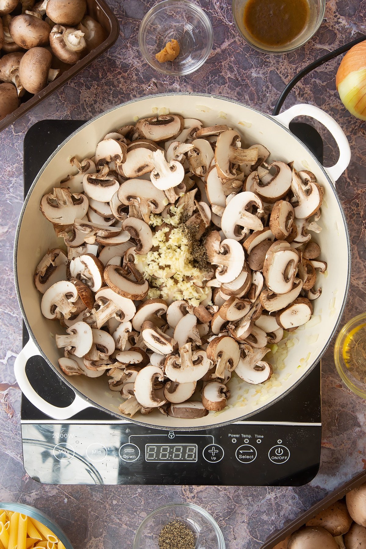 diced onions in a large white frying pan topped with garlic and sliced mushrooms on an induction hob.
