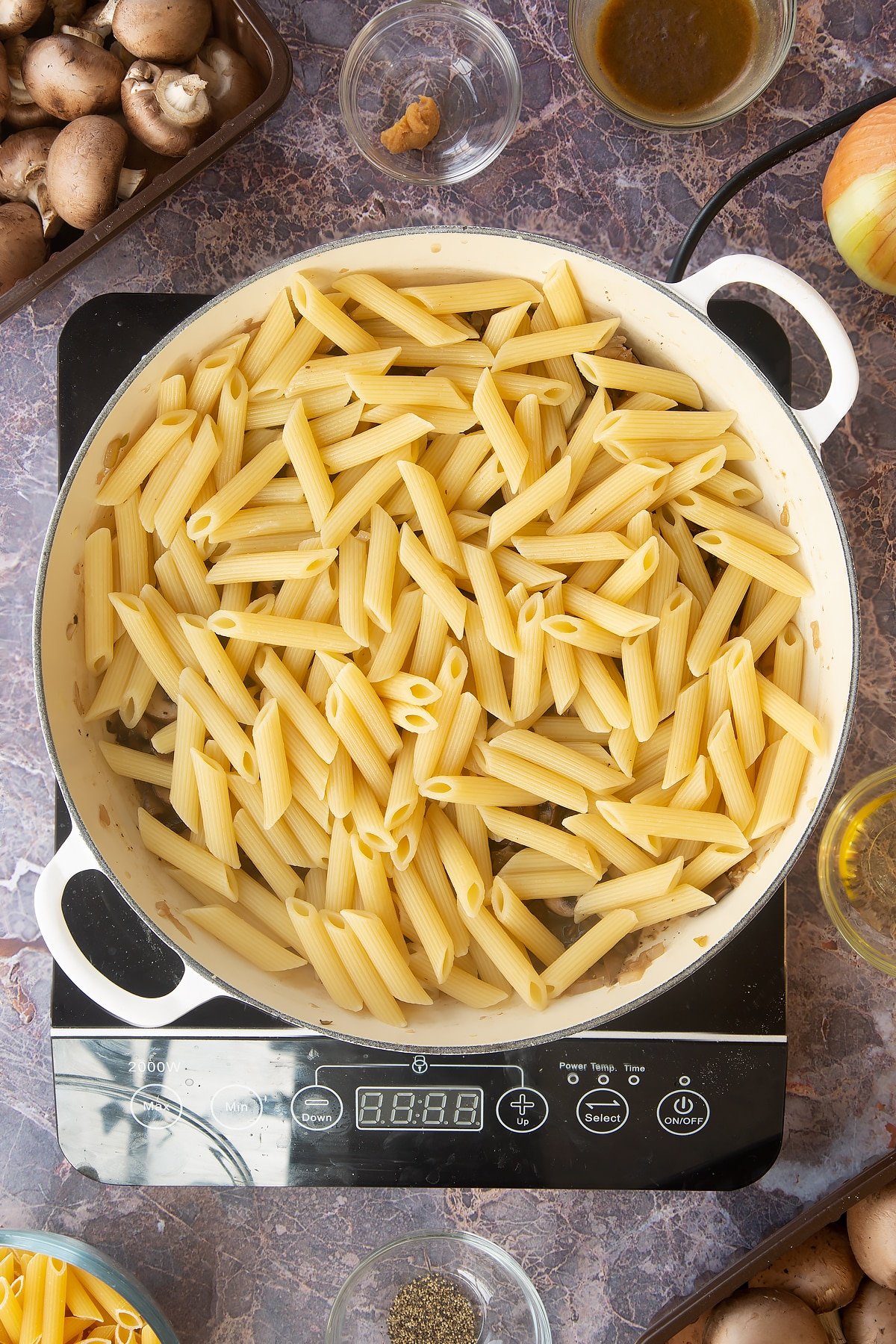 cooked mushroom, garlic and onion mix topped with cooked penne pasta in a large white frying pan on an induction hob.