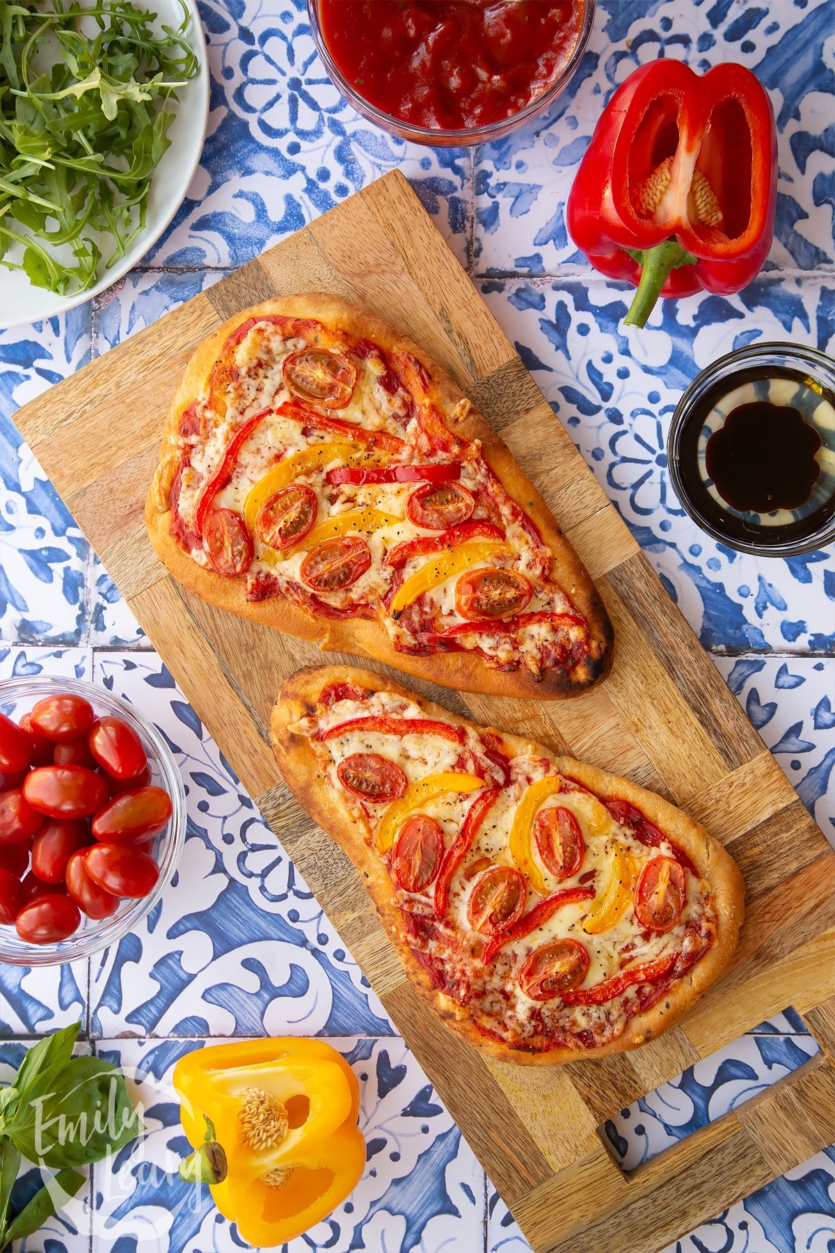 Two Naan bread pizzas on a wooden chopping board which is sat on top of some blue and white decorative tiles. Surrounding the Naan bread pizza are some of the ingredients required for the recipe.
