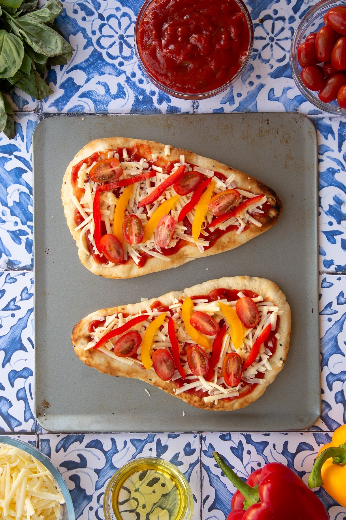 Two naan breads topped with tomatoes, grated cheese, sliced peppers and chopped tomatos sit on top of a square baking tray.