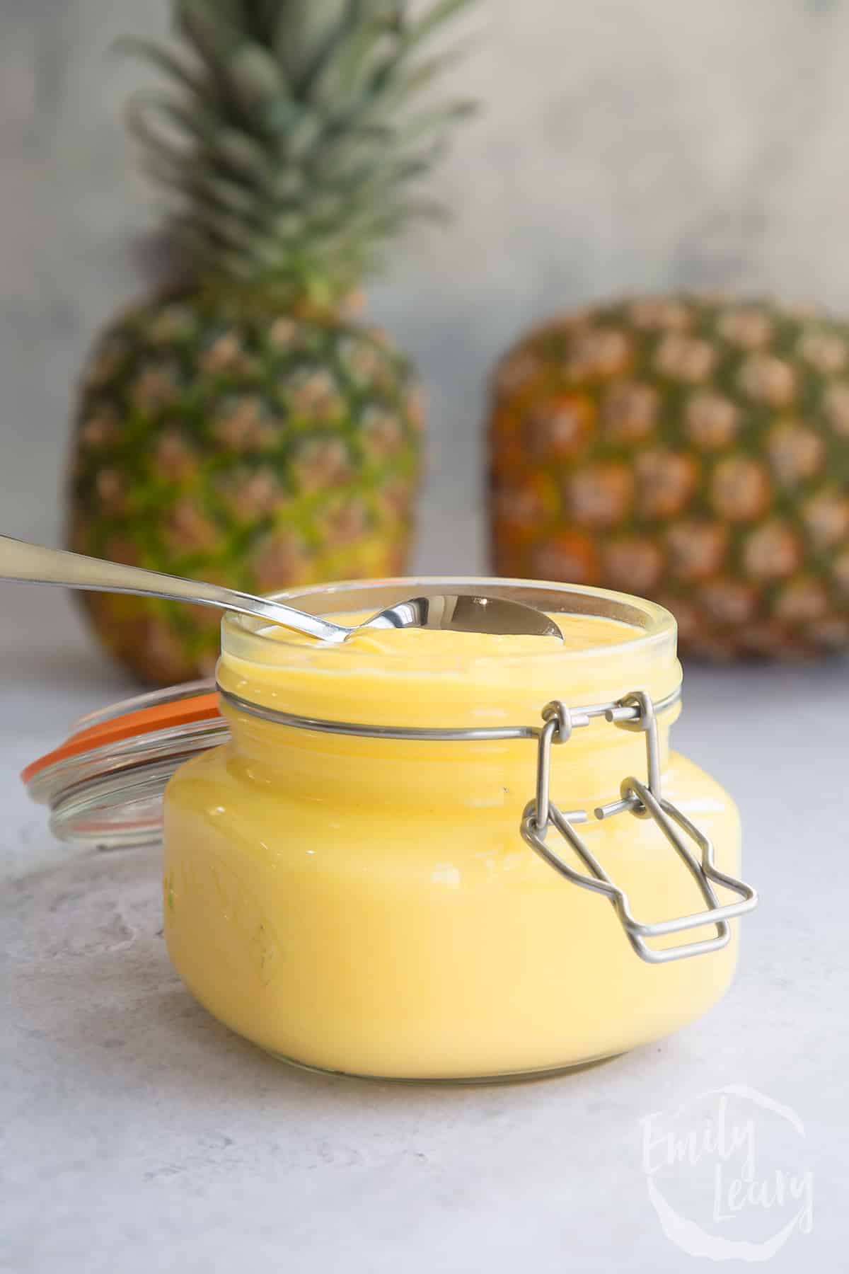 a jar filled with pineapple curd topped with a spoon with 2 pineapples in the background.