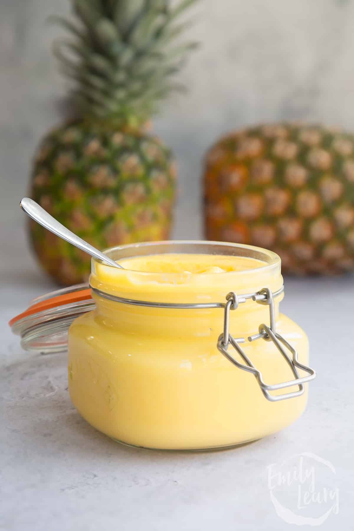 pineapple curd mixture in a glass jar with pineapples in the background and a spoon in the jar.