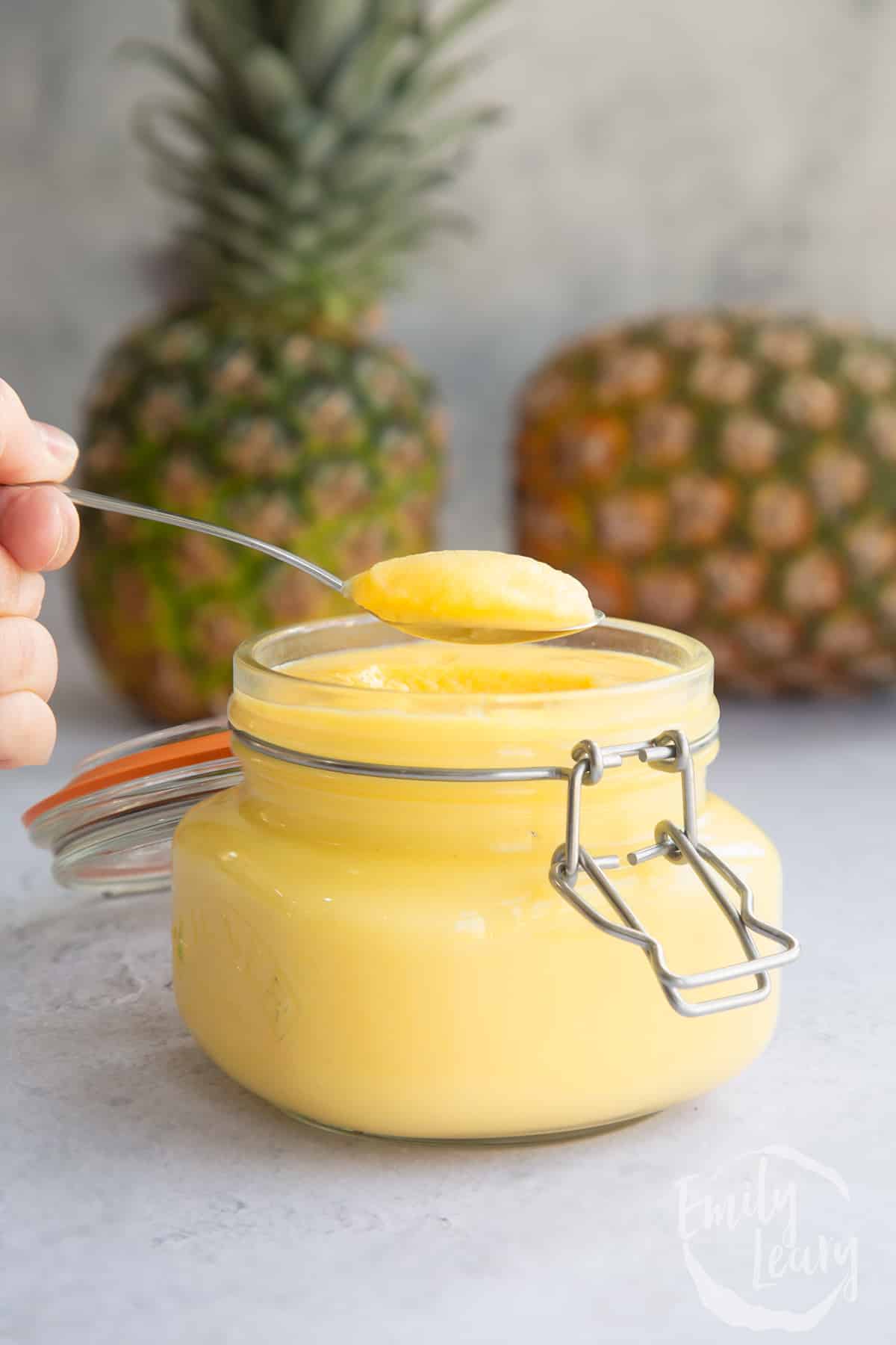a jar filled with pineapple curd topped with a spoon with 2 pineapples in the background.