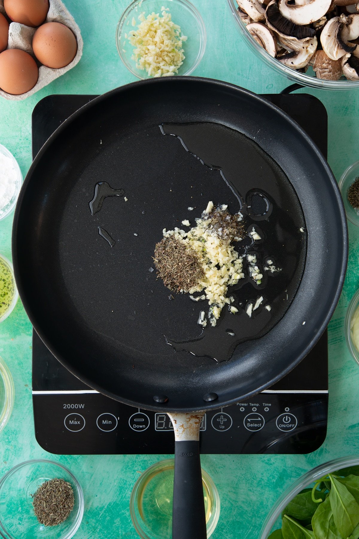 a large black frying pan on an induction hob with minced garlic, herbs and oil.