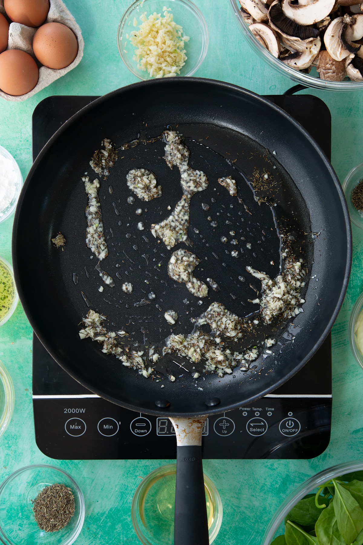a large black frying pan on an induction hob with mixed minced garlic, herbs and oil.
