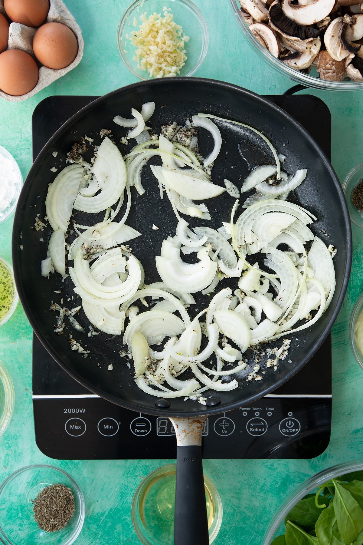 a large black frying pan on an induction hob with minced garlic, herbs and oil topped with uncooked onions.