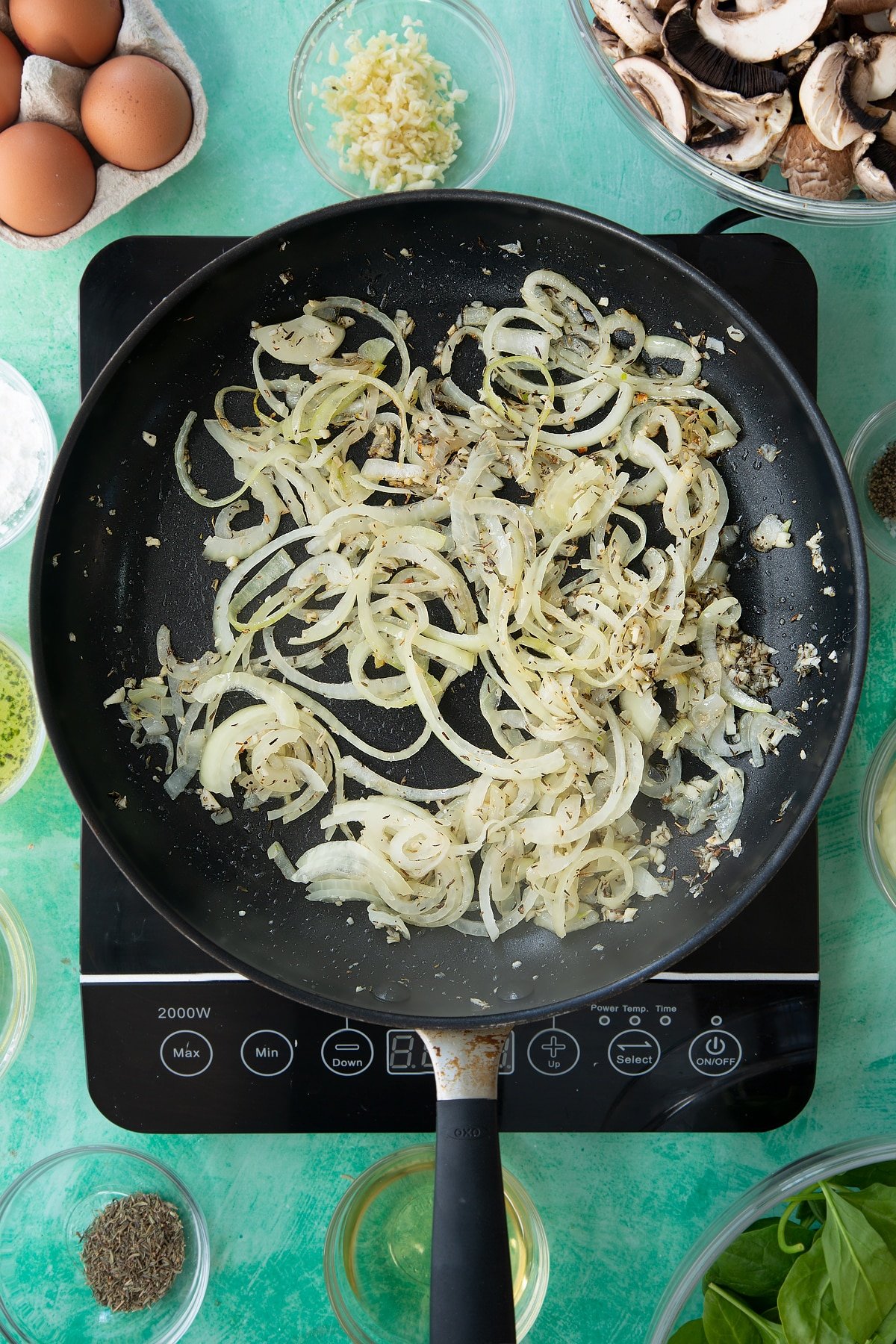 a large black frying pan on an induction hob with minced garlic, herbs and oil topped with cooked onion slices.