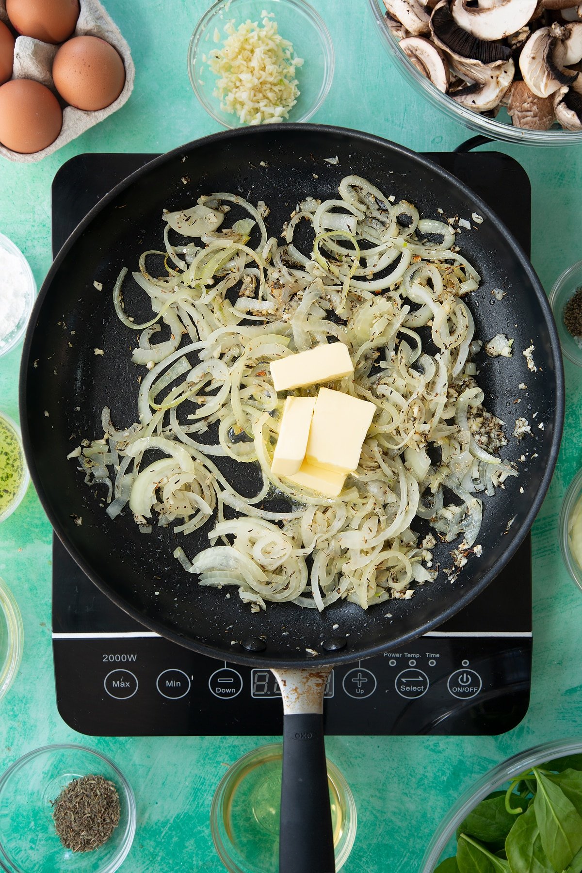 A large black frying pan with cooked onions, garlic and herbs topped with cubes of butter.