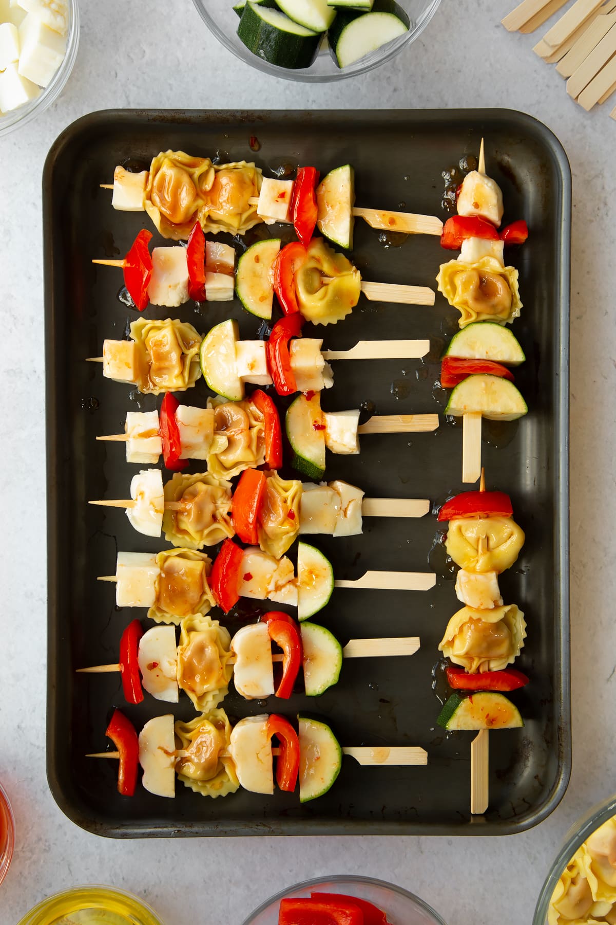 raw courgette, tortellini, halloumi and red pepper on wooden skewers placed on a baking tray drizzled in oil.