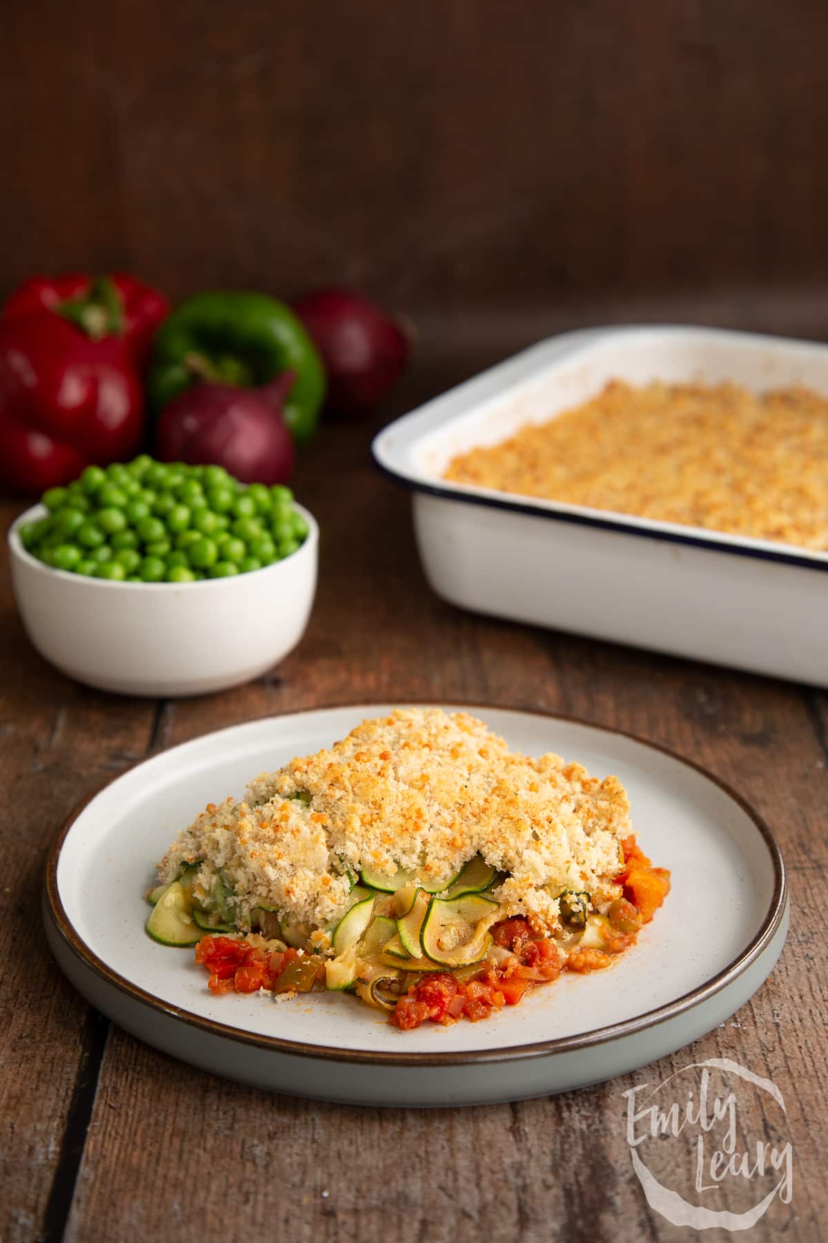 a piece of Tomato and courgette gratin on a white plate with a bowl of peas in the background.