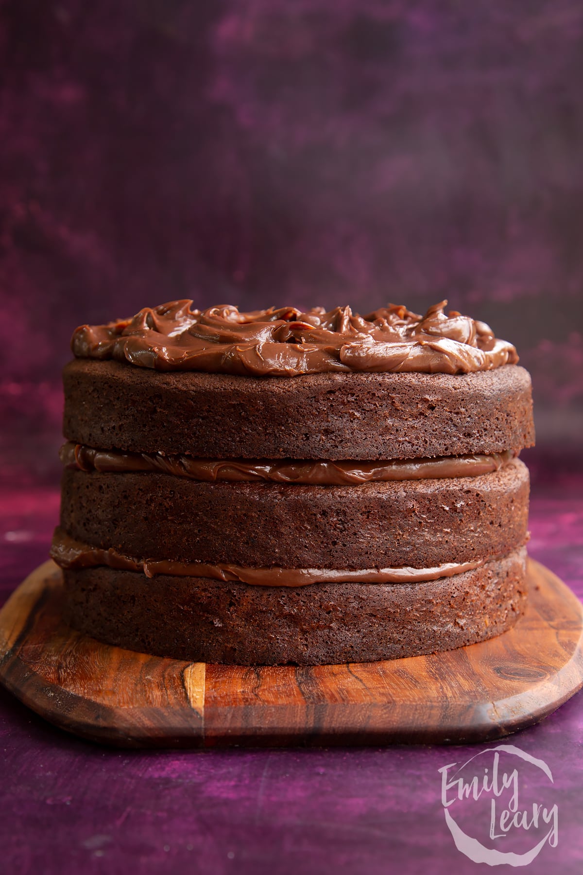 a side view of a 4 tier chocolate fudge cake on a wooden chopping board.