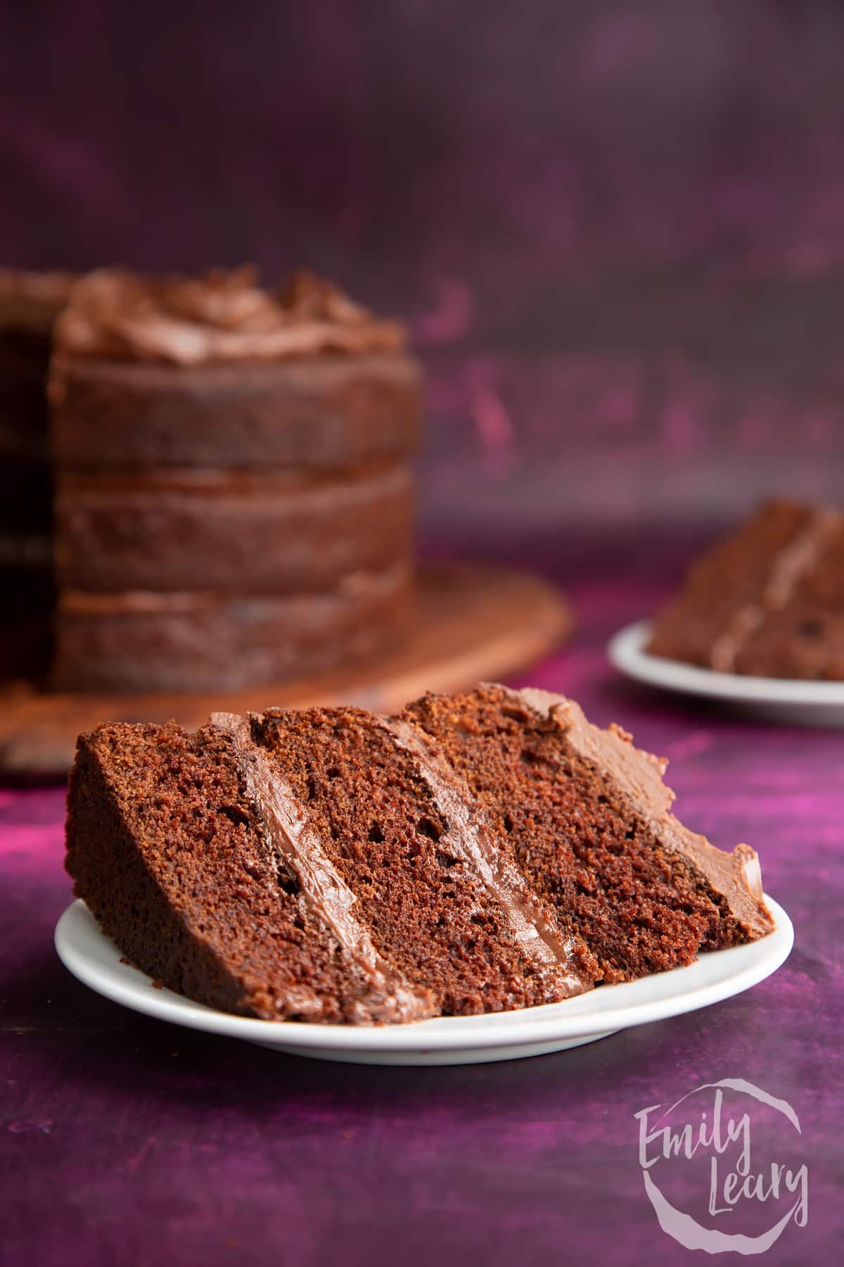 a slice of chocolate fudge cake on a white plate with the full cake in the background.