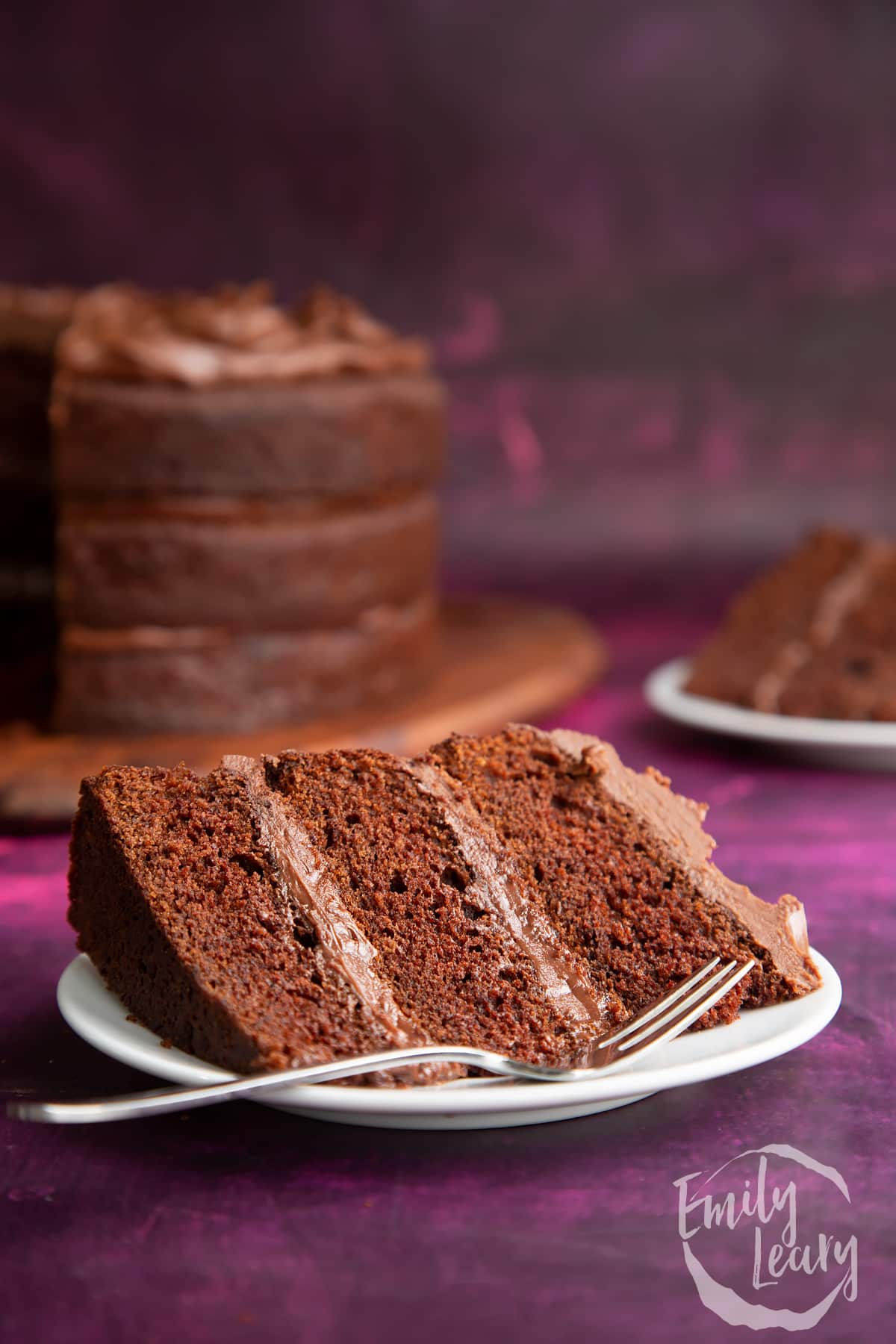 a slice of chocolate fudge cake on a white plate with a fork at the front.