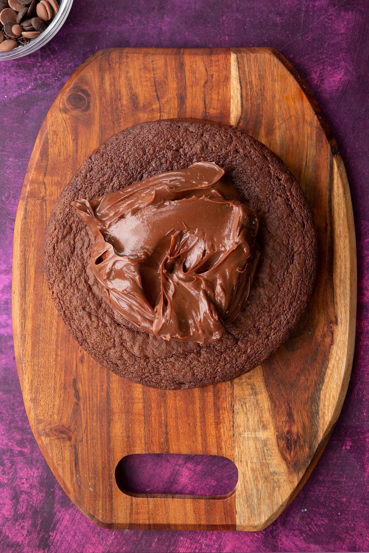 a large round chocolate sponge cake on a wooden chopping board topped with chocolate fudge.