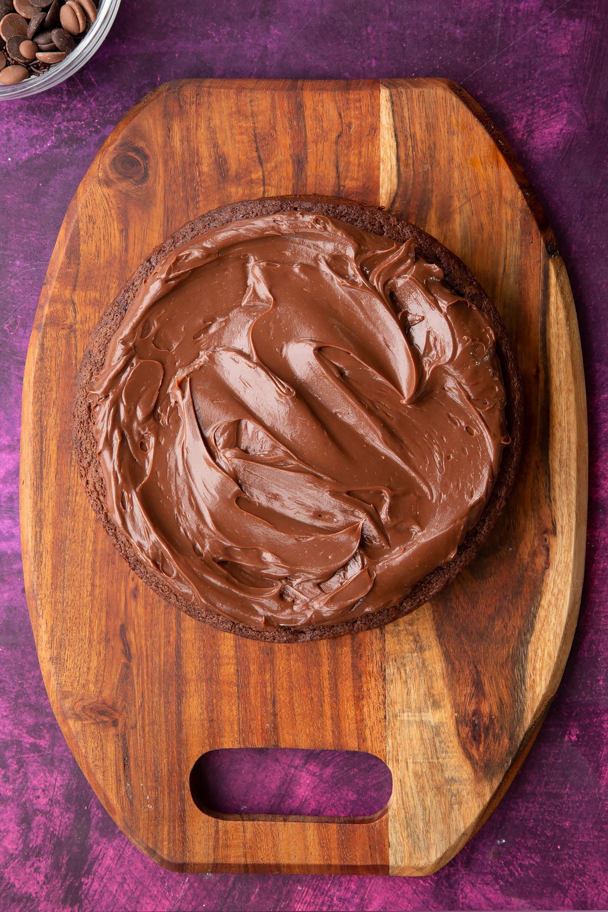 a large round chocolate sponge cake on a wooden chopping board topped with chocolate fudge and spread evenly.