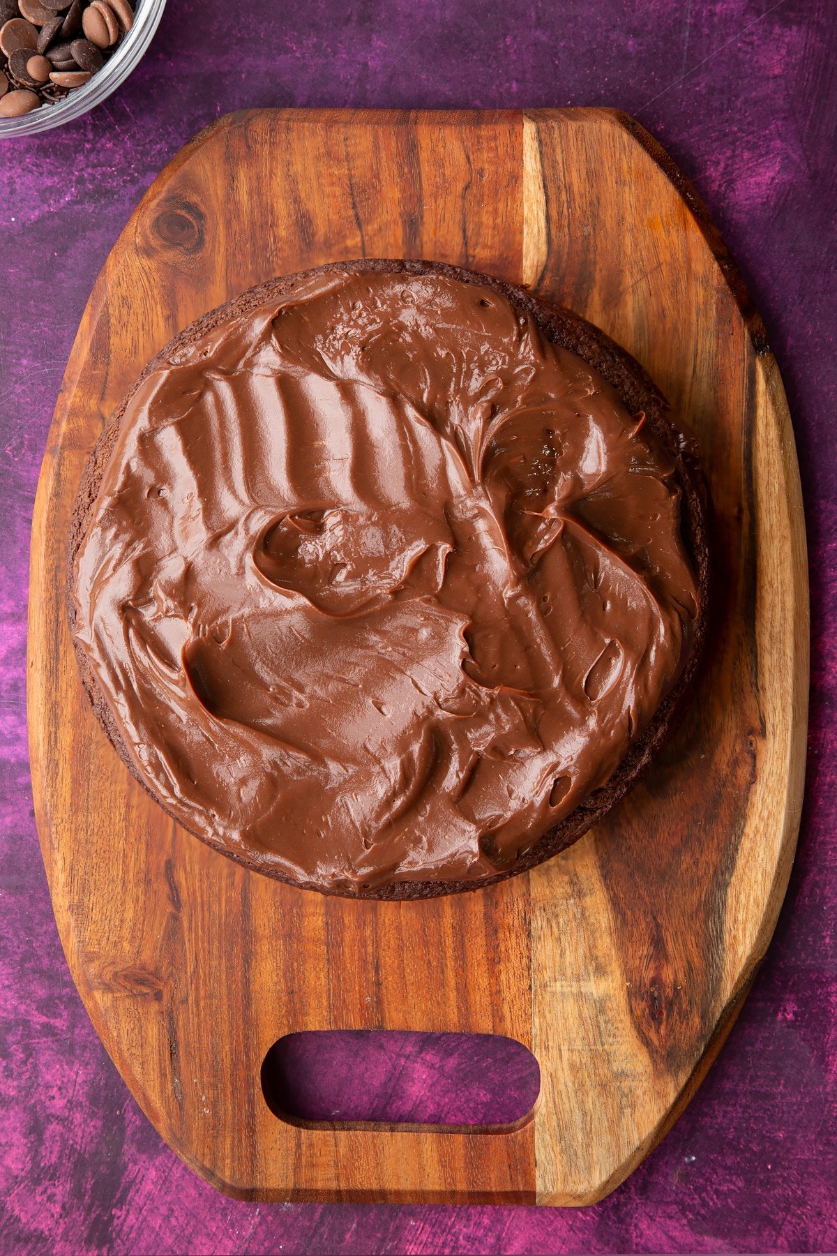 a large round chocolate sponge cake on a wooden chopping board topped with chocolate fudge and spread evenly.