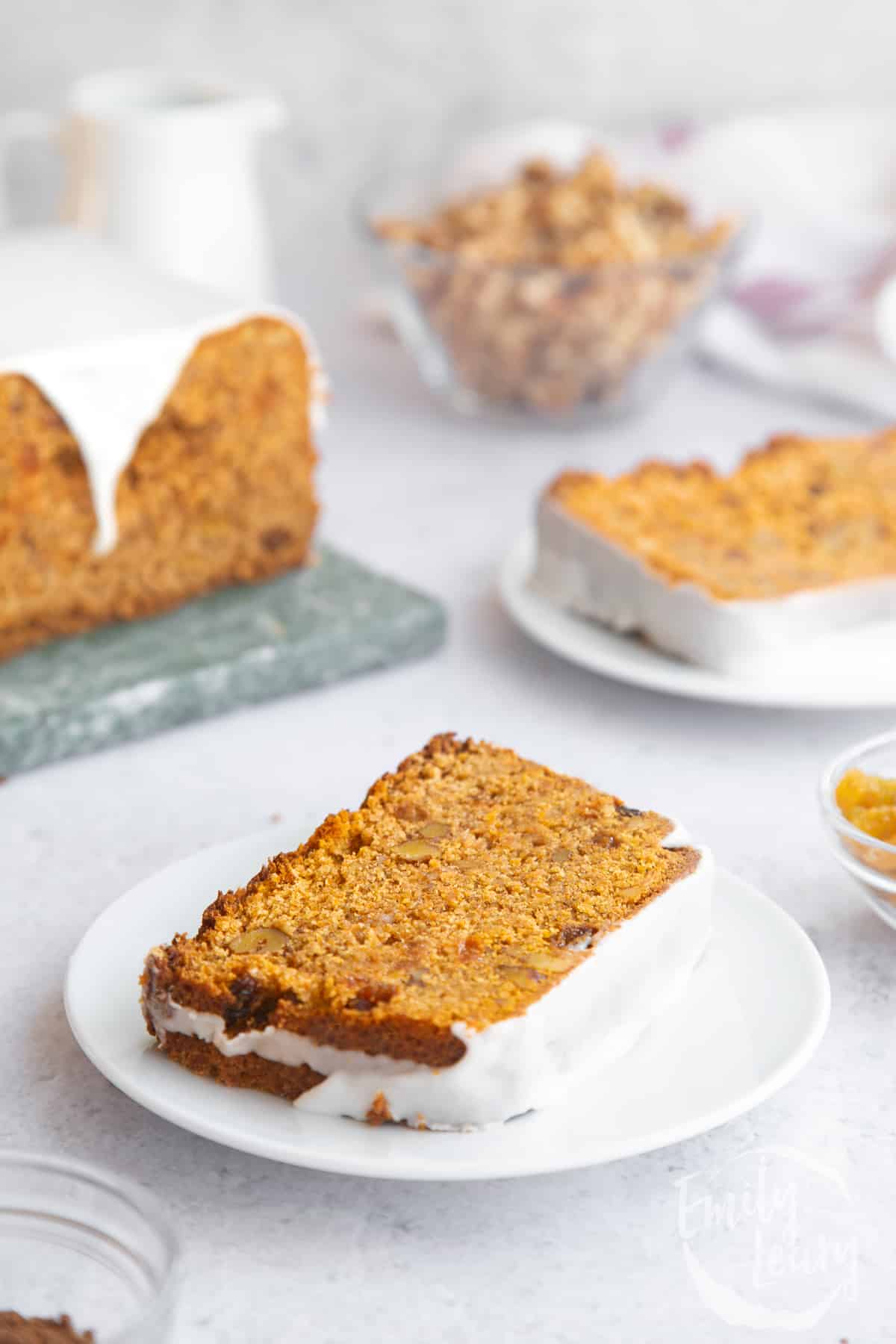 a slice of Vegan carrot loaf cake on a white plate with the full loaf in the background.