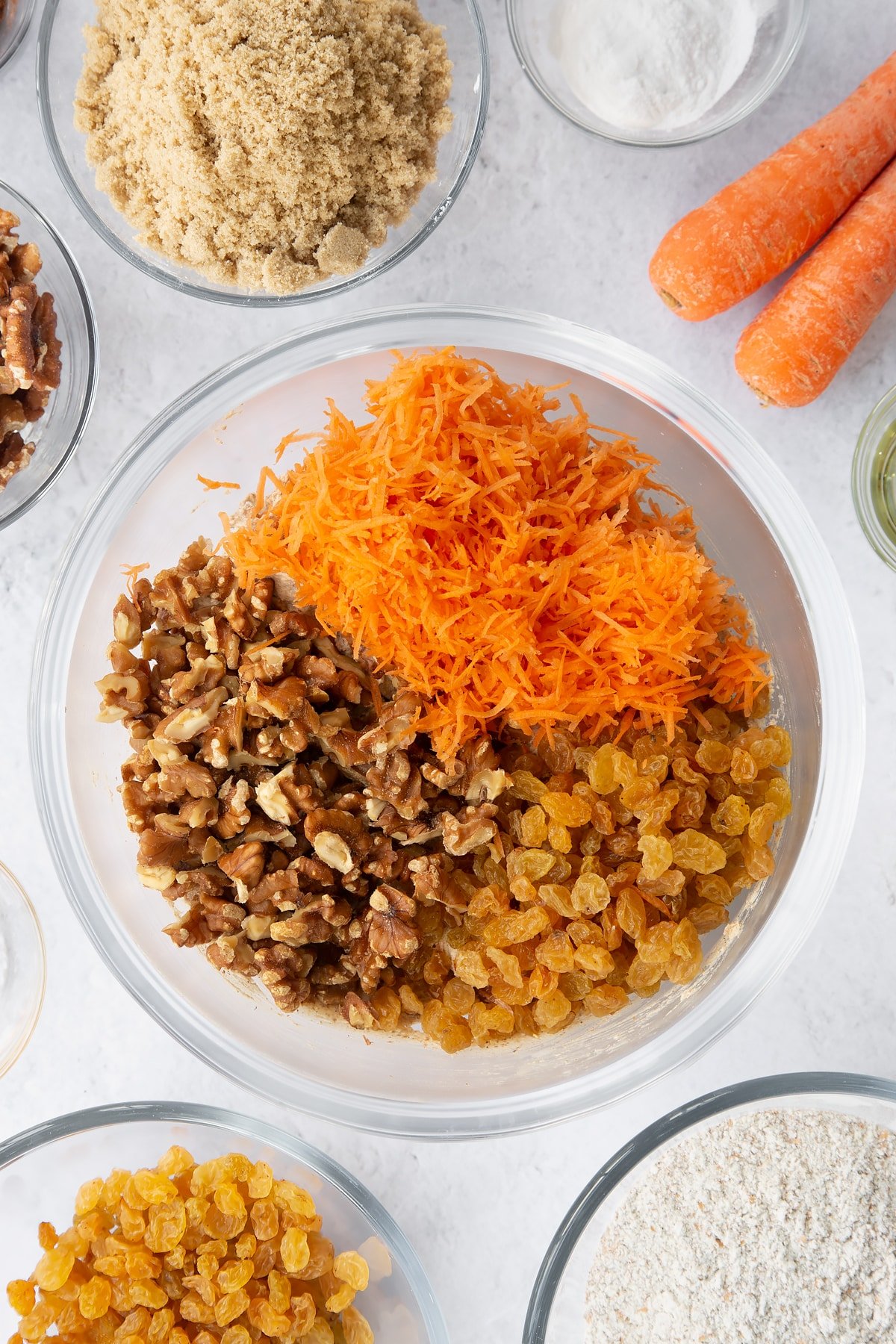 mixed Vegan carrot loaf cake batter in a large clear bowl topped with sultanas, walnuts and grated carrot.