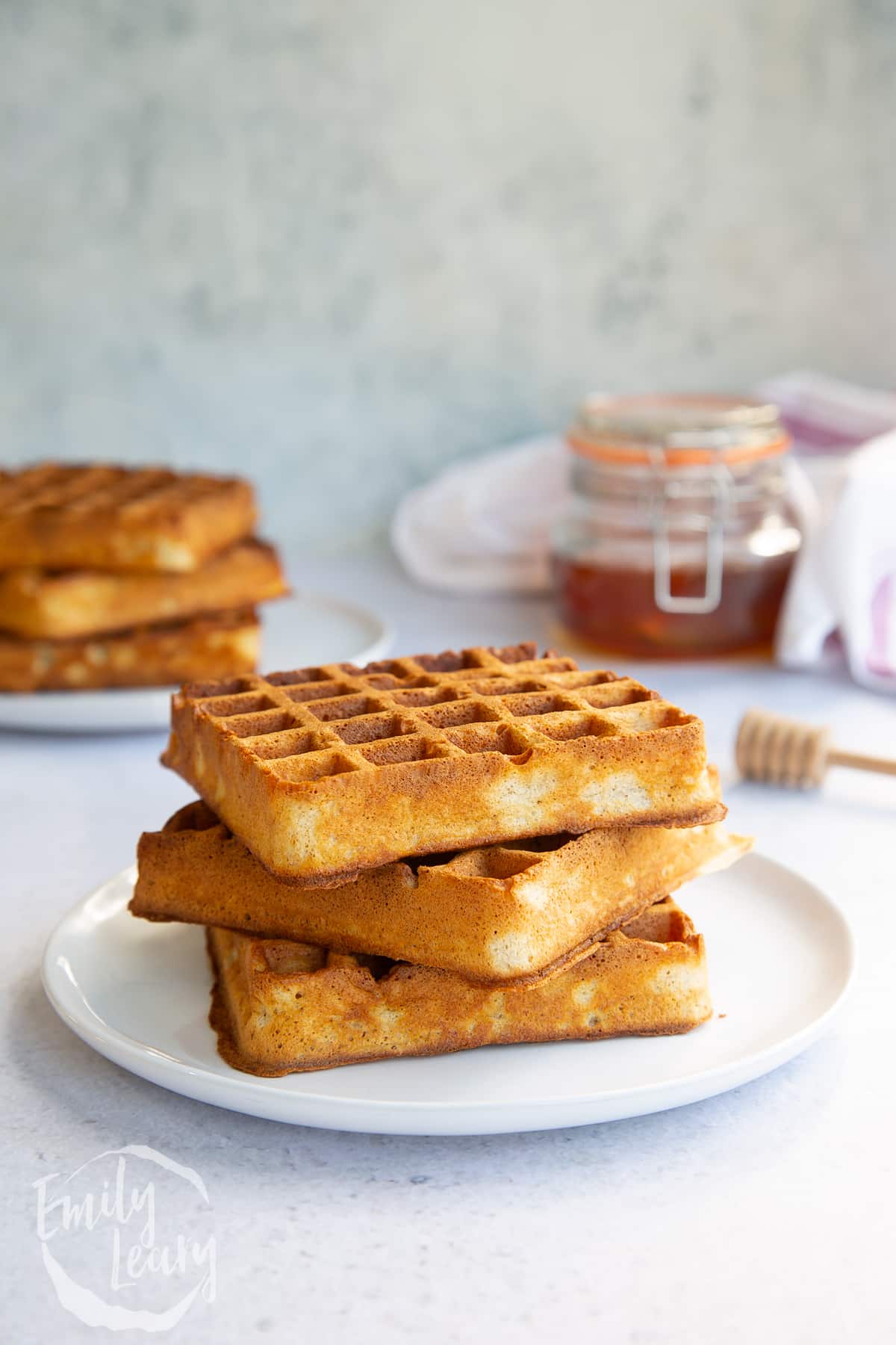 3 wholmeal waffles stacked on a white plate with honey and more waffles in the background.