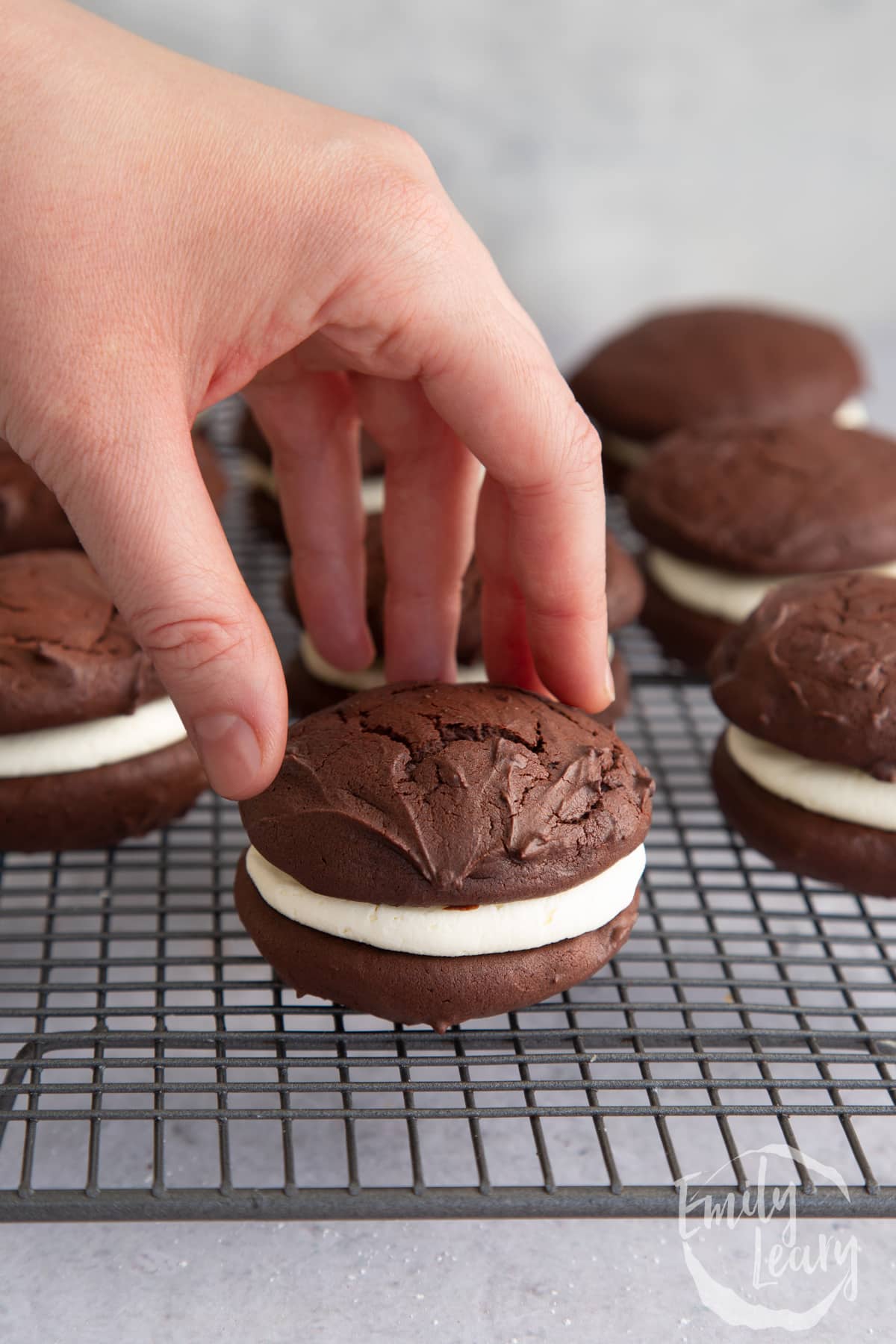 a hand holding a chocolate whoopie pie on a wire cooling rack with more in the background.