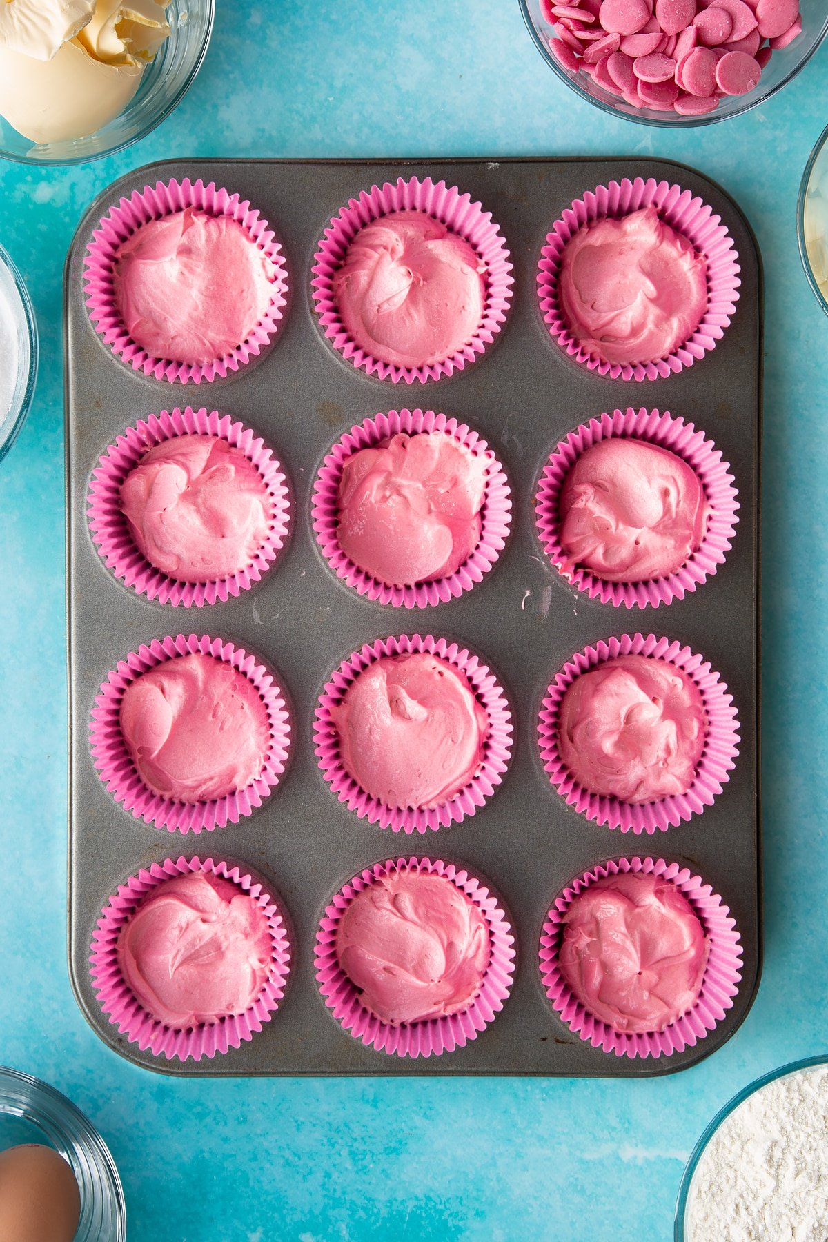 12 pink cupcake liners filled with pink cupcake mix in a cupcake tray.