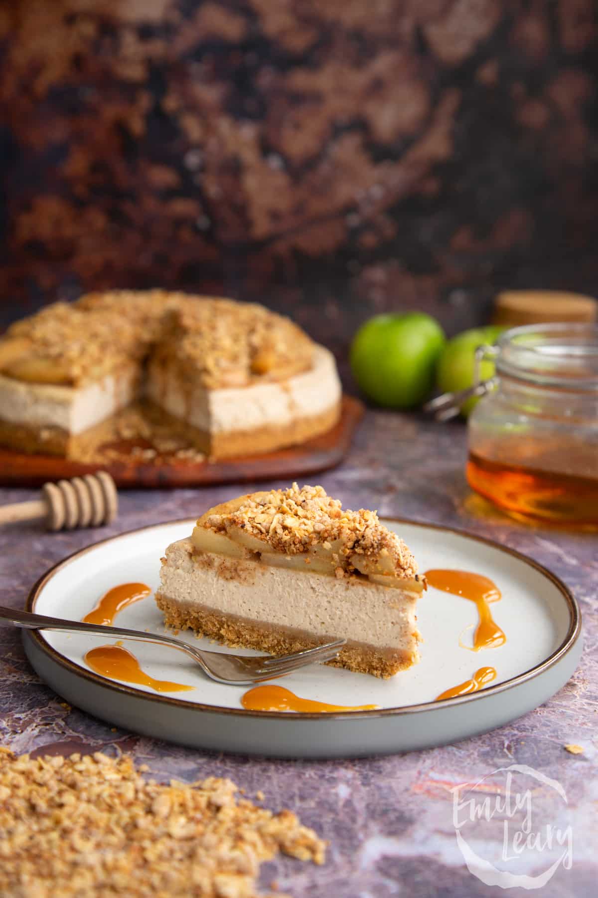 A finished slice of apple crumble cheesecake with a fork on the side, served on a white decorative plate. 