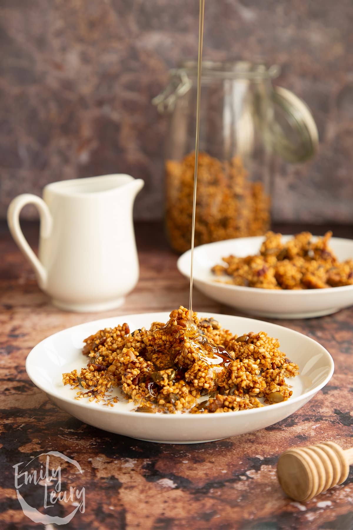 Steel cut oats granola in a white bowl next to a white milk jug with honey drizzled on top.